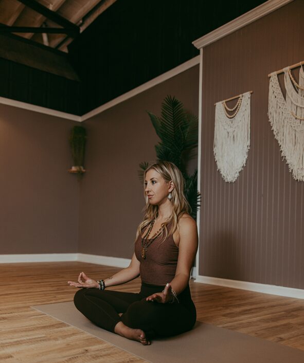 Woman sitting cross-legged on a yoga mat in a room with wooden floor and wall decorations, meditating peacefully.
