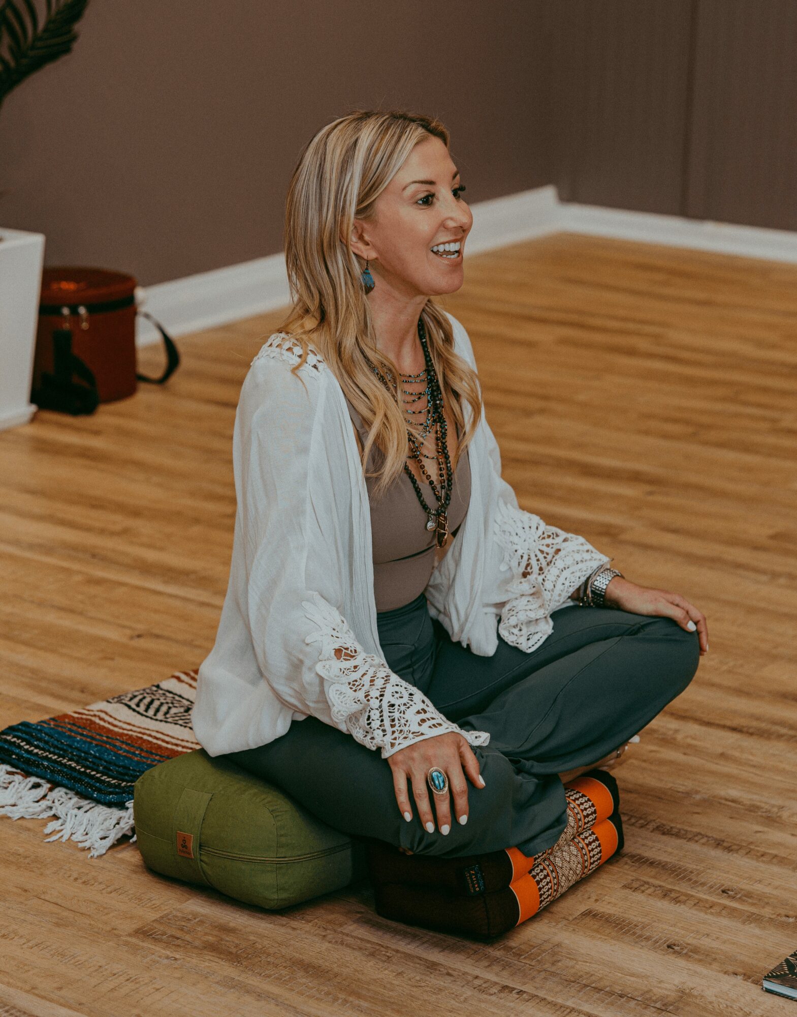 Woman sitting cross-legged on a cushion, smiling, in a room with wooden floor and brown wall.