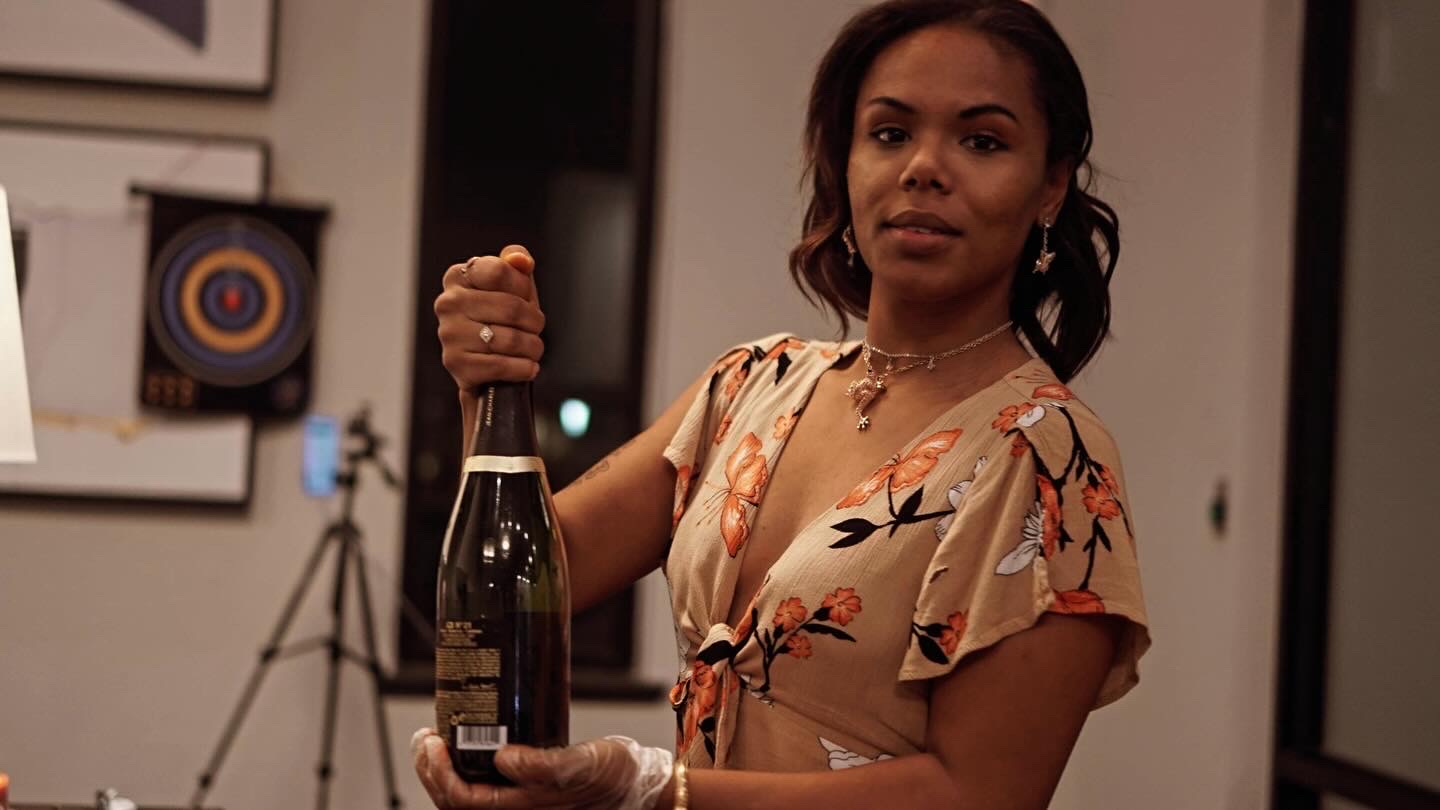 Woman holding a bottle in a room with a dartboard and tripod in the background.