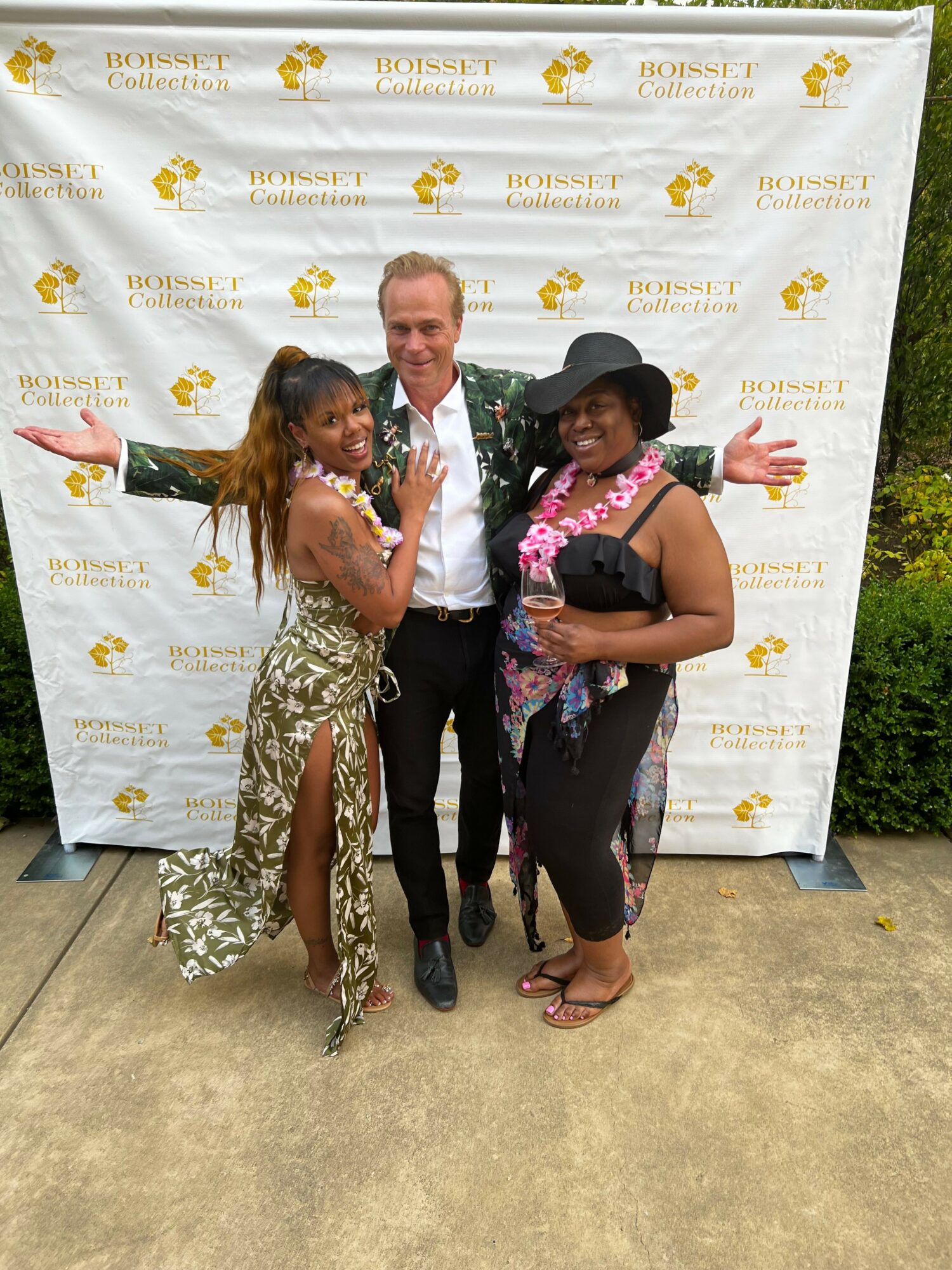 Three people posing in front of a white backdrop with yellow logos, one woman in a floral dress, one man in a white shirt, one woman in a black hat and floral top.