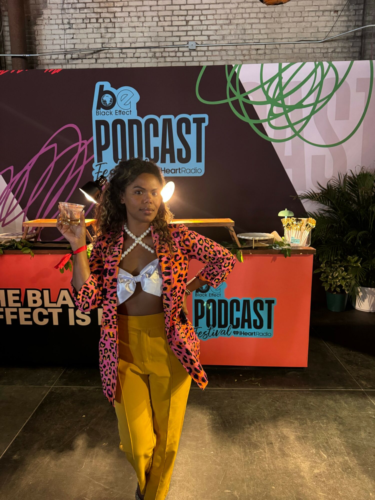 Person standing at a podcast event, holding a glass, with a backdrop and table behind them.