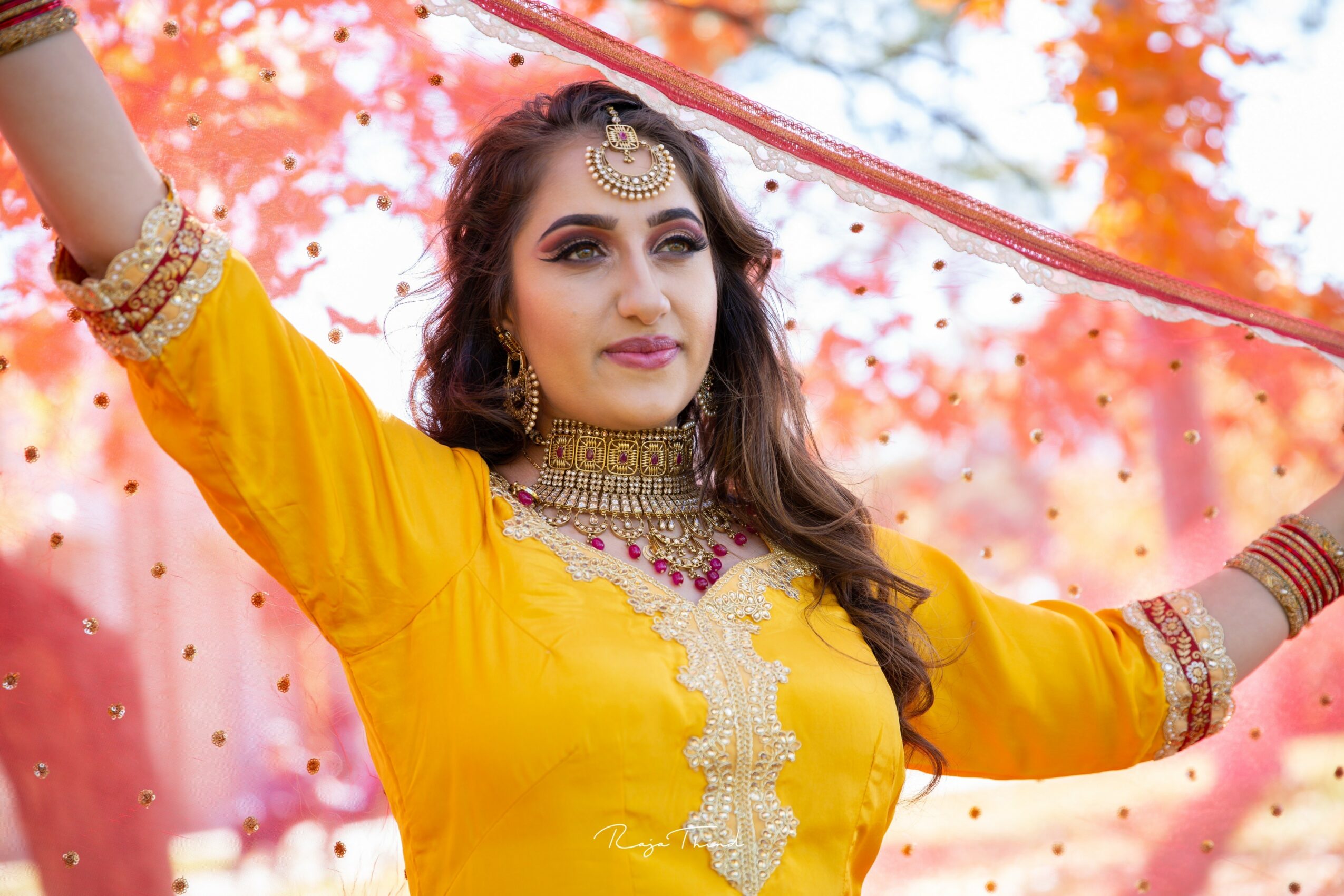 Woman in yellow traditional attire holding a red veil outdoors with autumn trees in background.