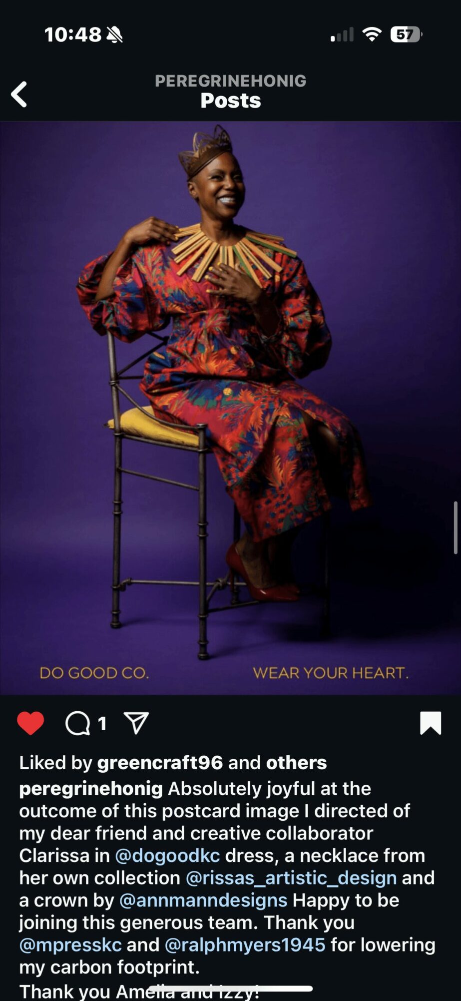 Woman sitting on a chair, smiling, wearing a colorful dress and jewelry, against a purple background.