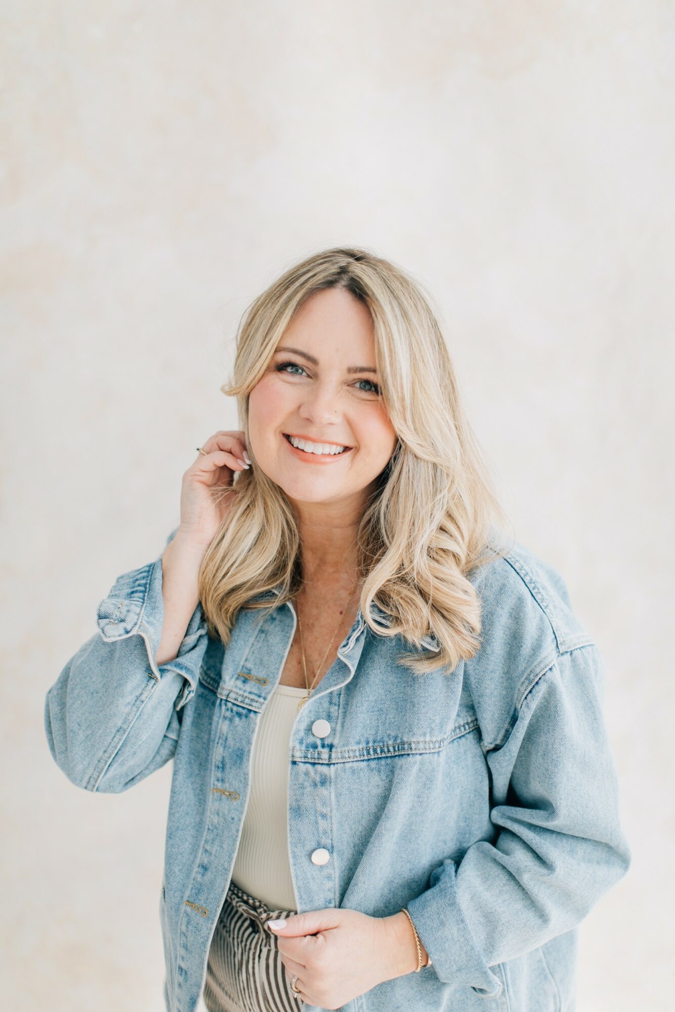 Smiling woman with blonde hair wearing a denim jacket, touching her hair, against a light background.