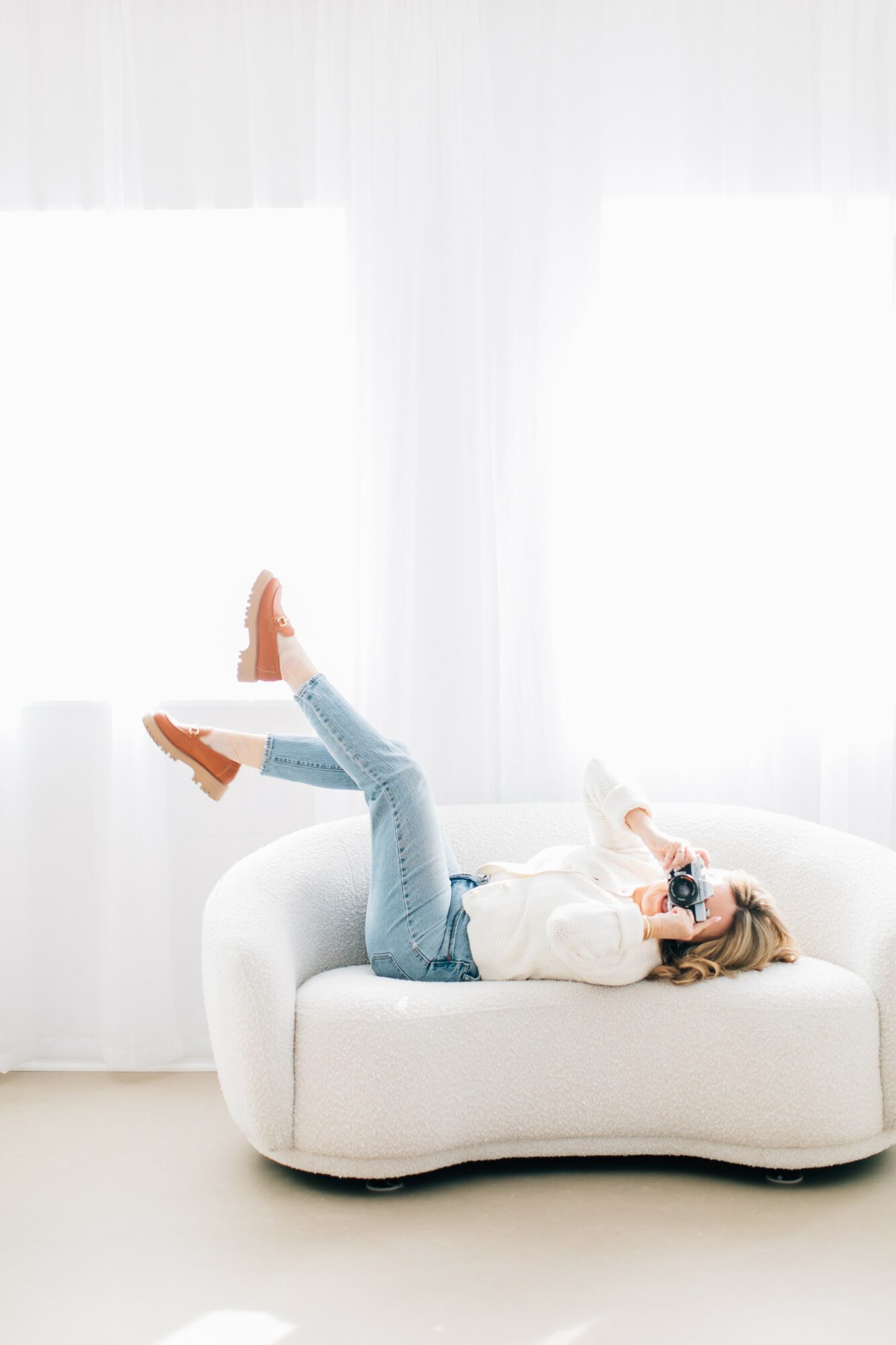 Person lying on a white sofa, wearing sunglasses, with legs raised, in a bright room with white curtains