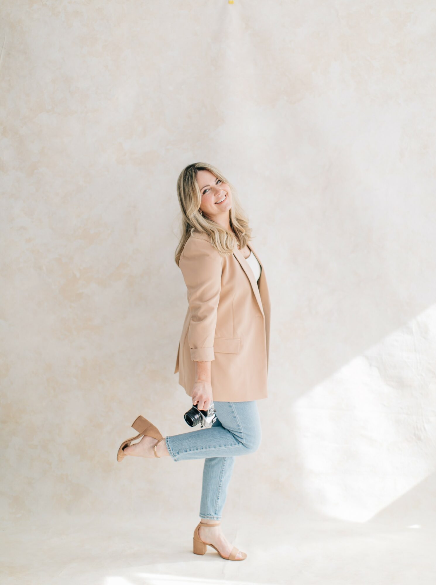 Young woman with blonde hair and glasses, wearing a beige coat, jeans, and heels, standing against a plain wall.