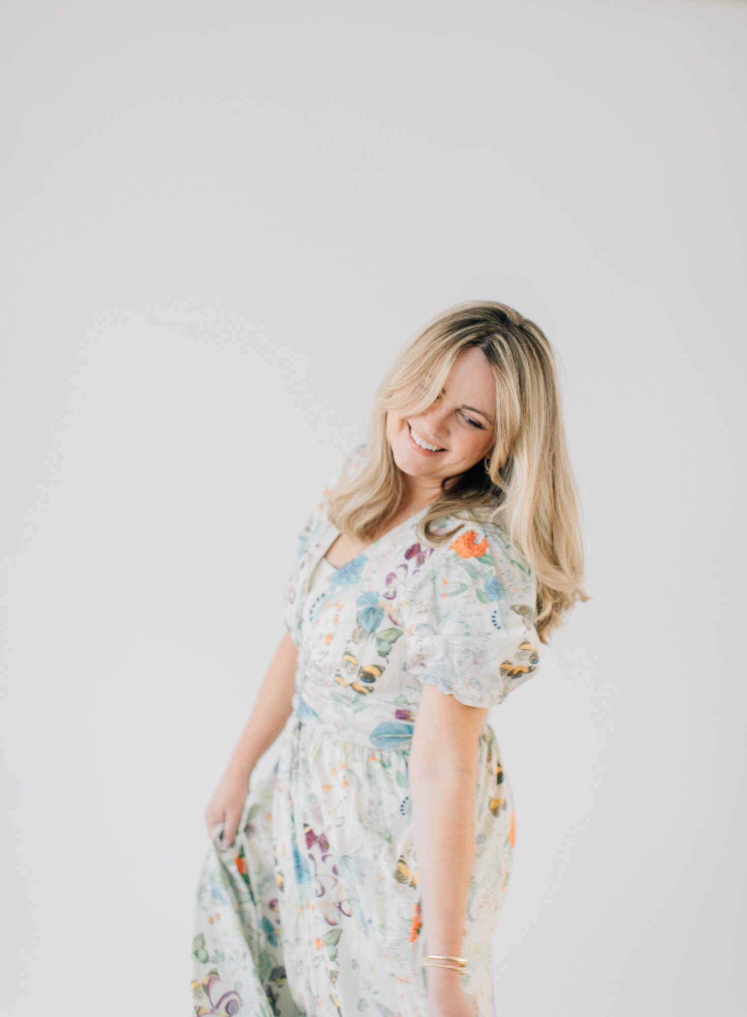 Woman with blonde hair wearing a floral dress, smiling and looking down, against a plain background.