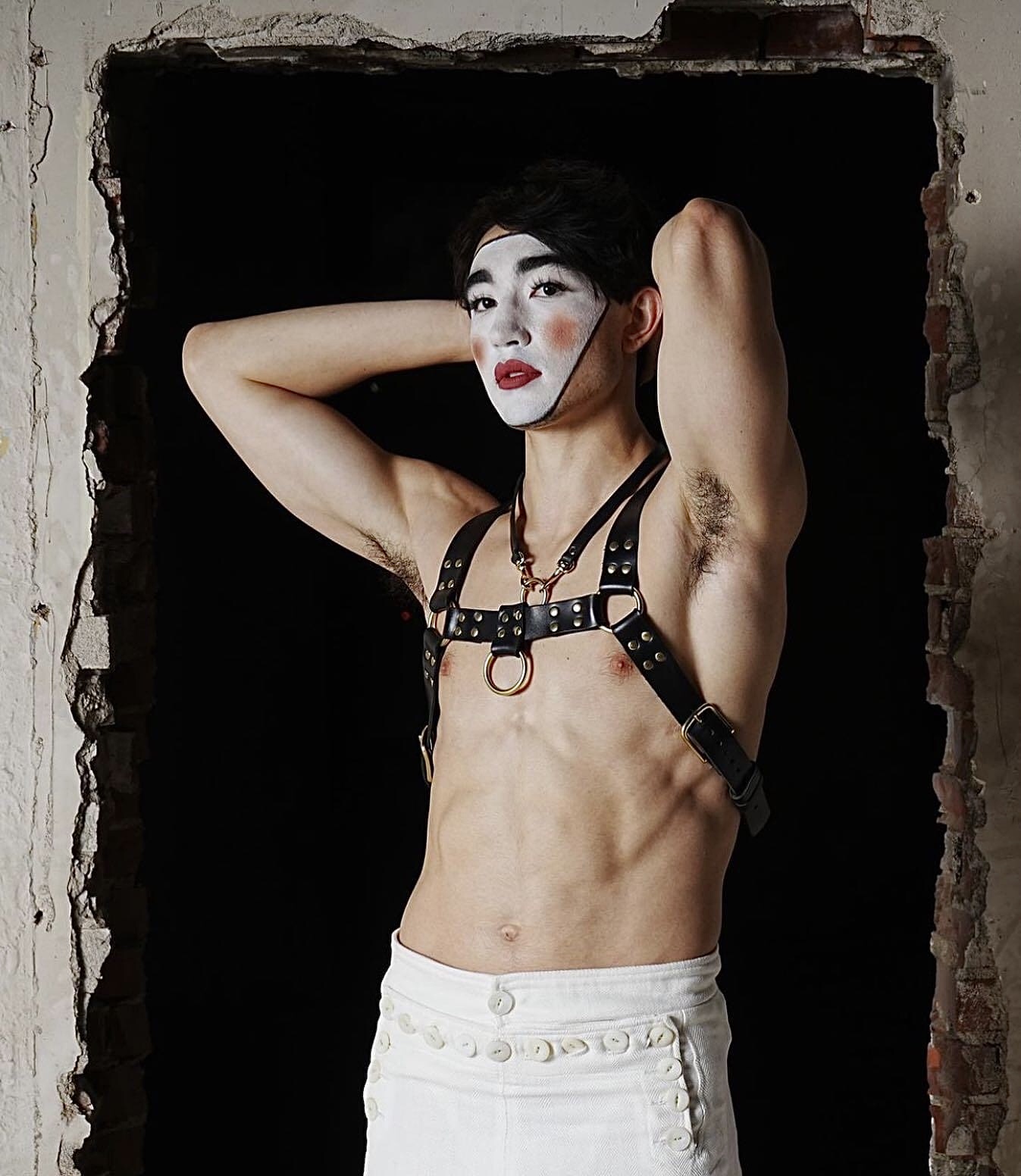 Person with makeup and harness posing with arms behind head, standing in a brick frame opening, dark background.