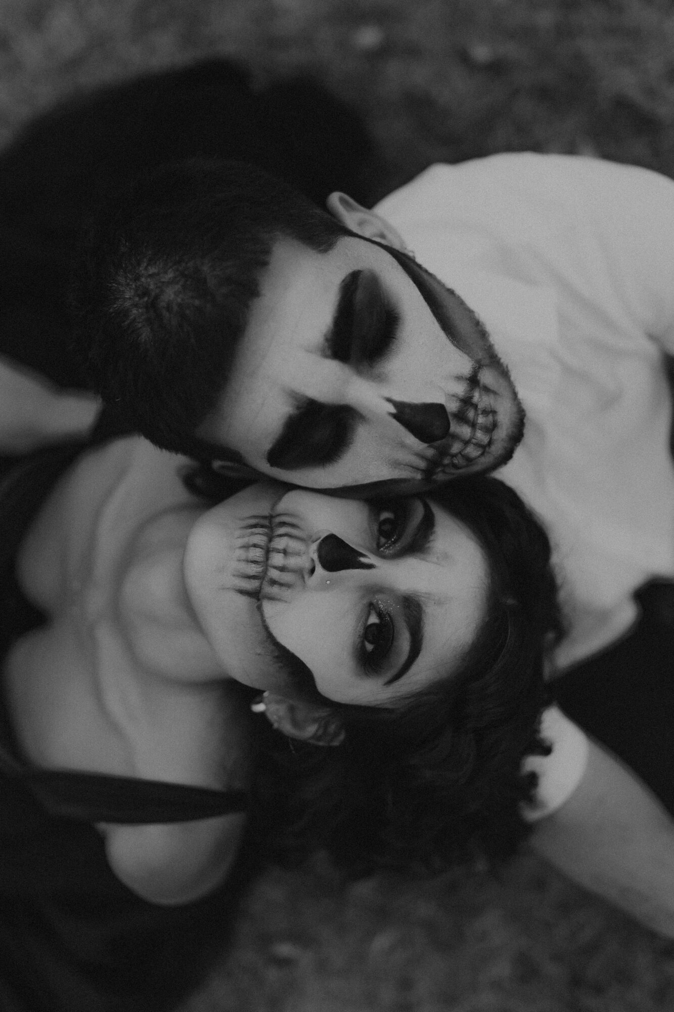 Two people with skull face paint lying close together, one with dark hair and the other with lighter hair, in black and white.