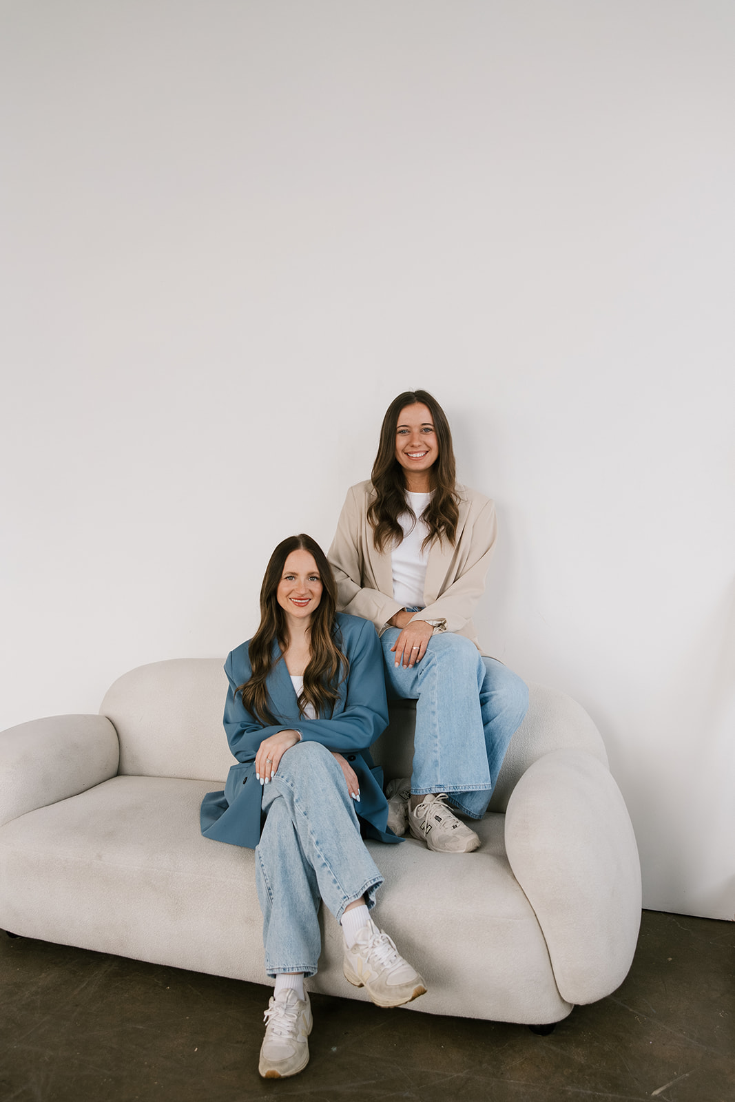 Two women with long hair, one sitting on a sofa and the other standing behind, smiling in a minimal room.