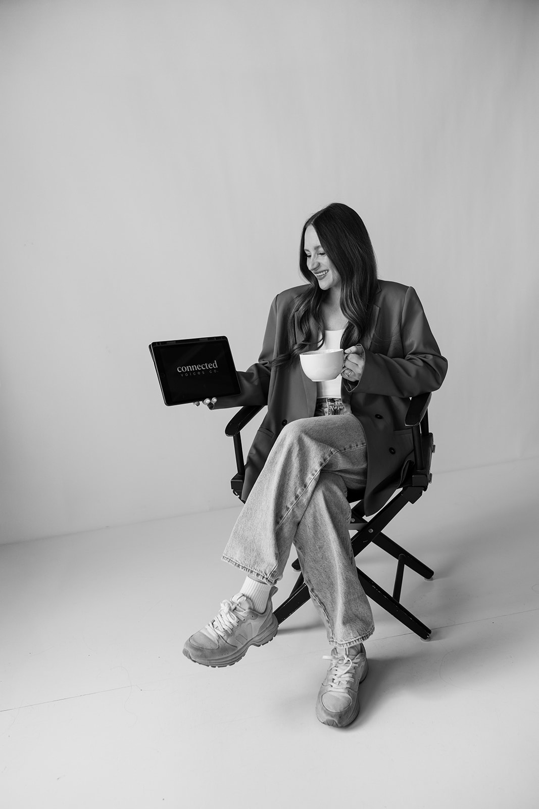 Woman sitting on a chair holding a mug and a laptop, wearing casual clothes, in a plain setting.