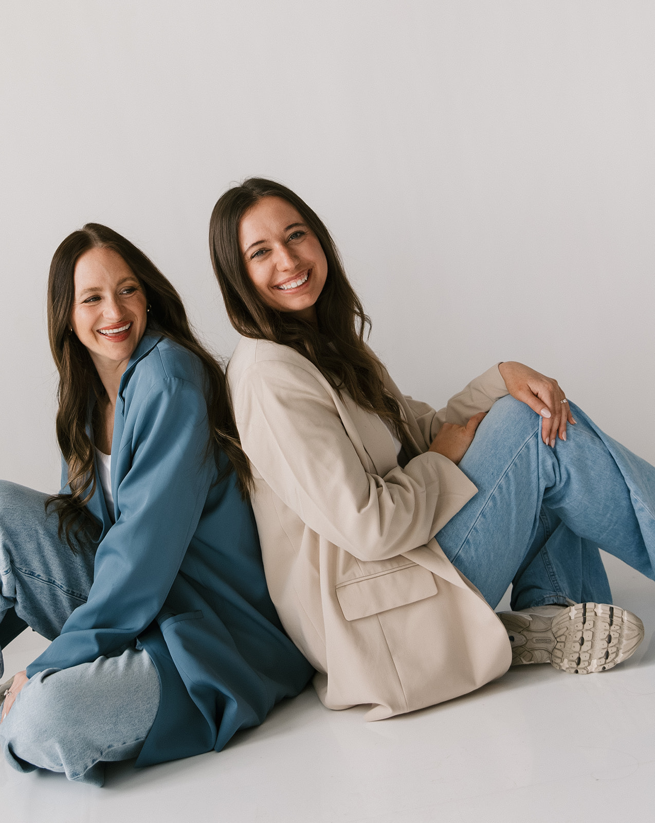 Two women sitting on the floor, smiling, against a plain white background.