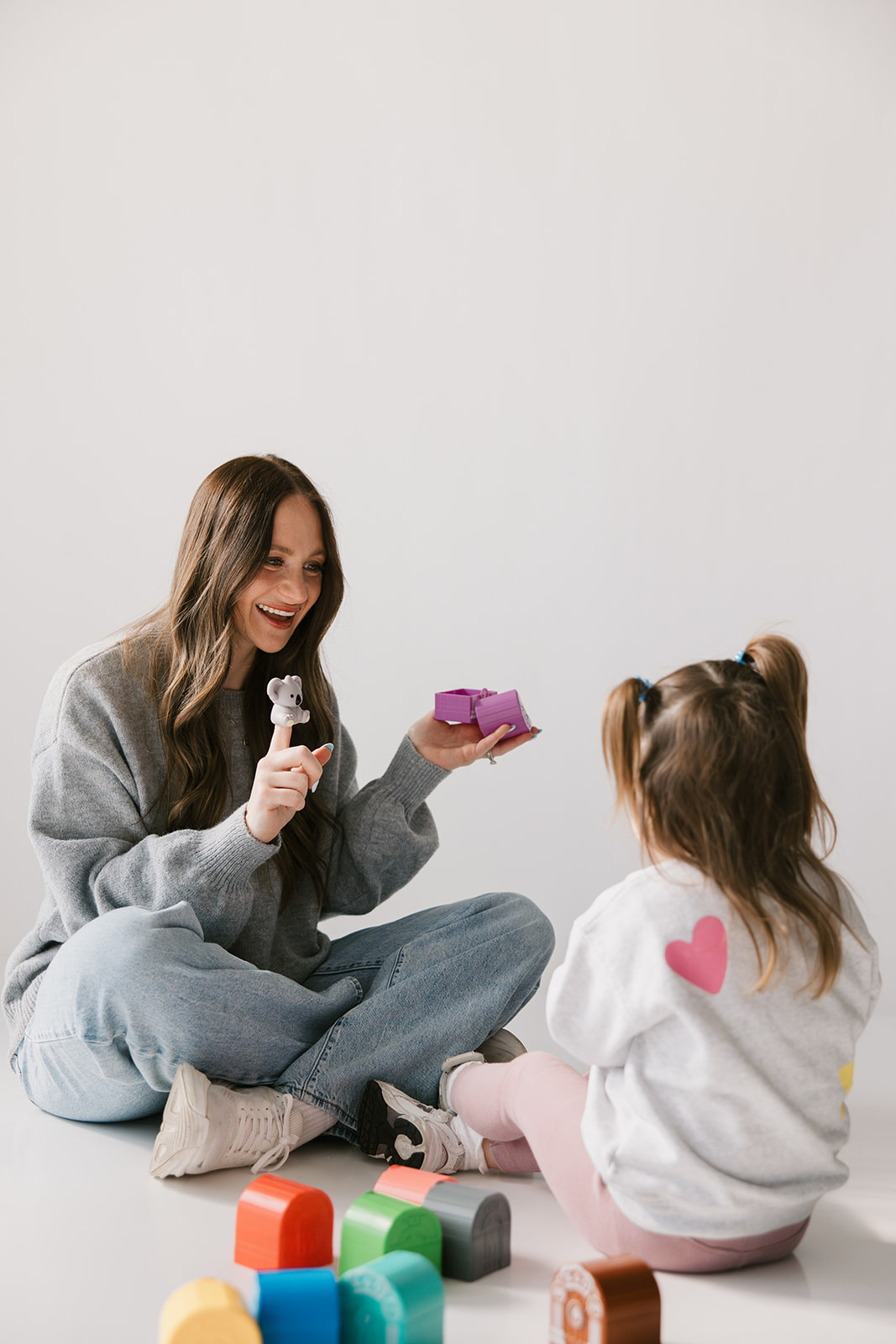 Two girls sitting on the floor, one holding a small gift box and a toy, smiling at each other, with colorful blocks nearby.