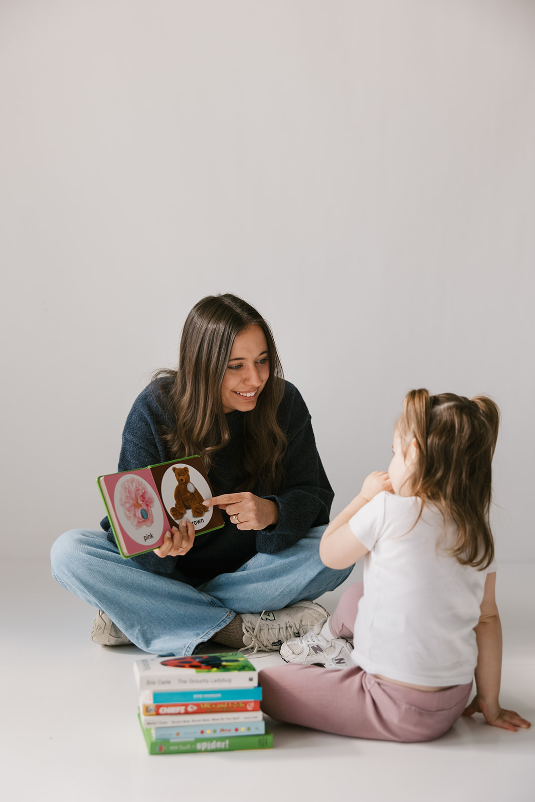 Adult woman reading a book to a young girl sitting on the floor with a stack of books nearby.