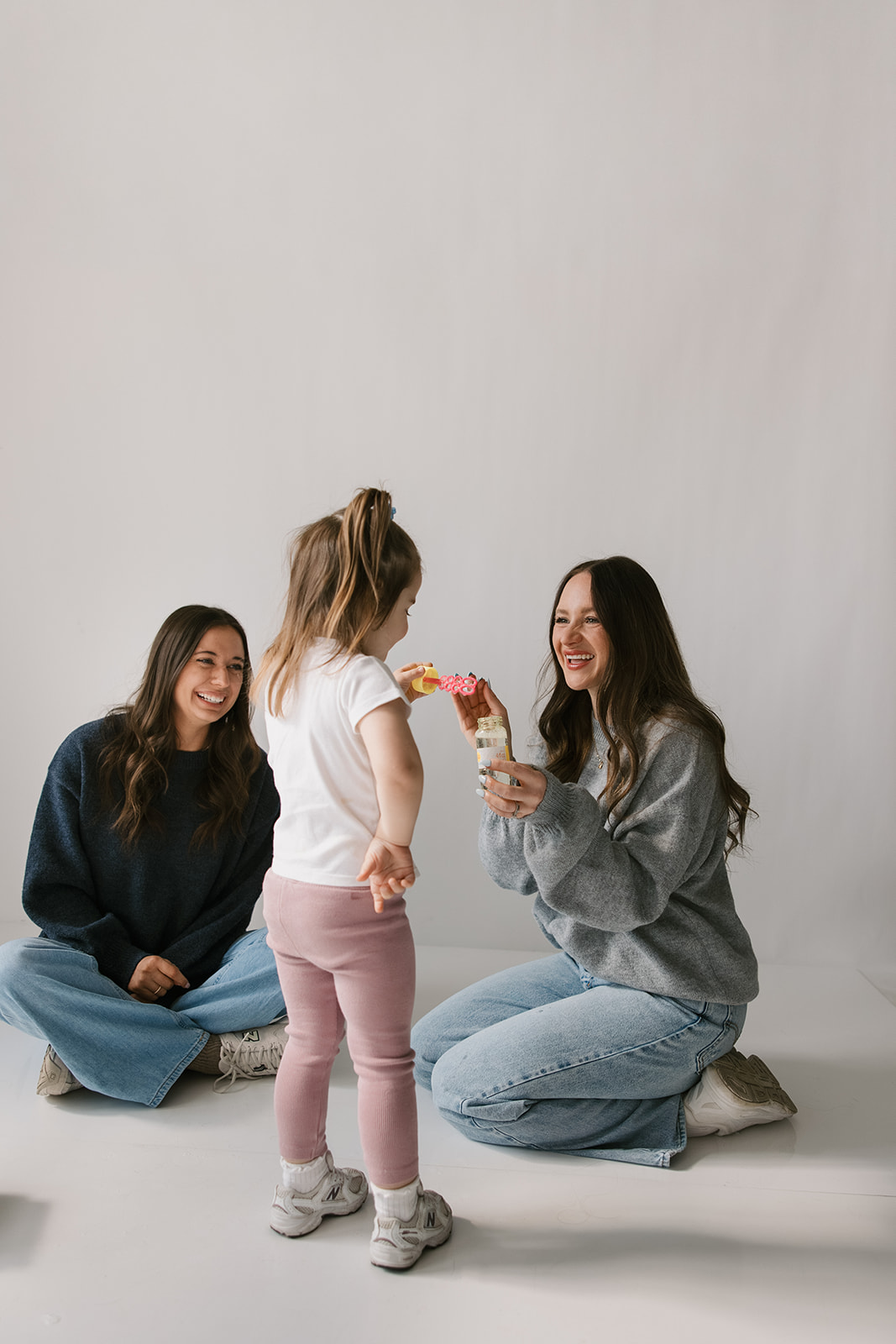 Three people, two women and a girl, sitting and standing on a plain white background, smiling and interacting.