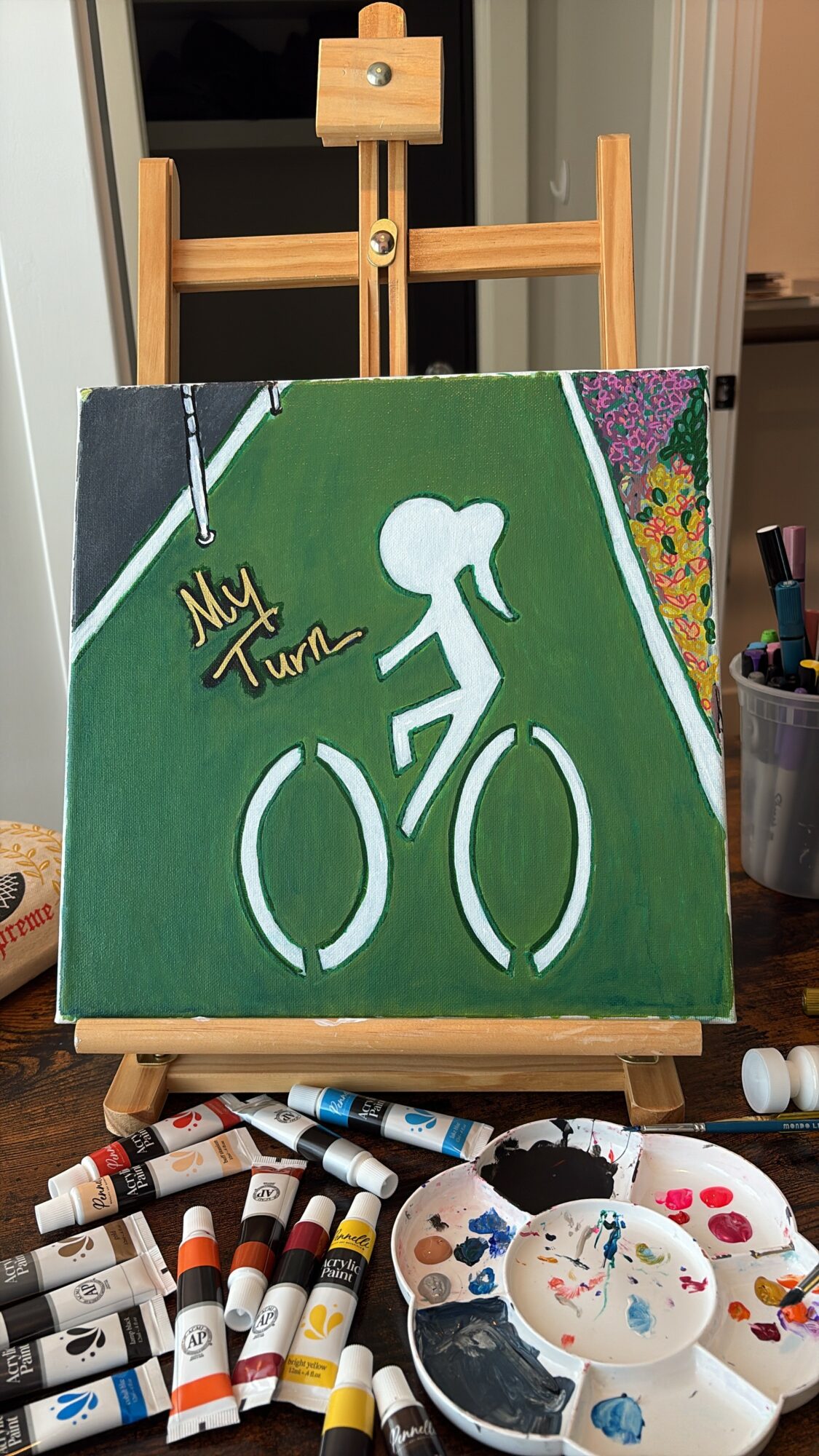 Painting of a person riding a bicycle on a green background with colorful flowers and a white path, on an easel.