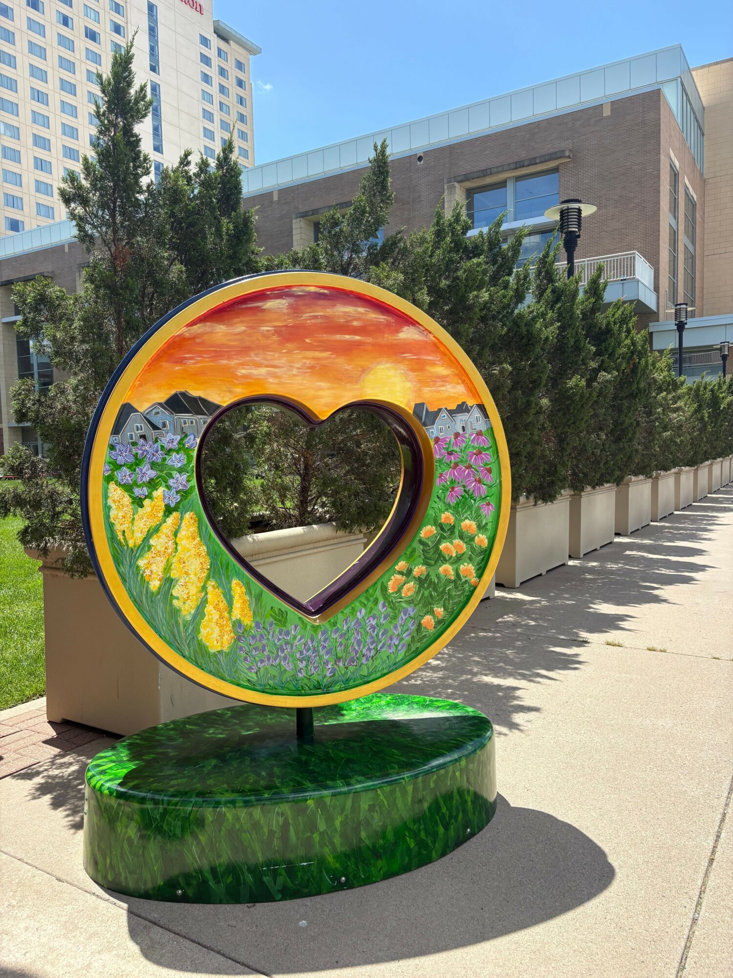 Colorful heart-shaped sculpture with sunset, flowers, and greenery, outdoors on a sunny day.