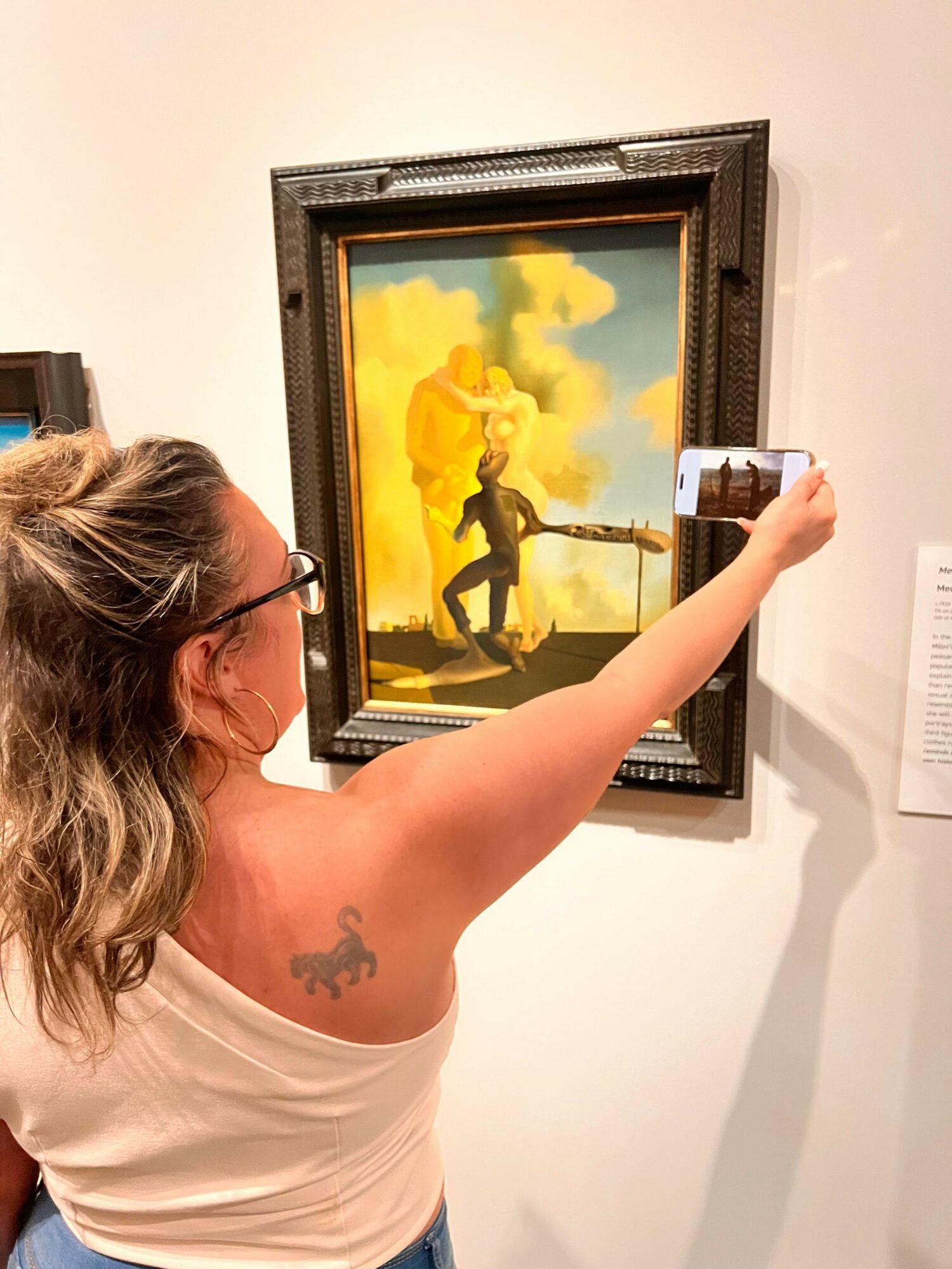 Person taking a photo of a painting with a phone, seen from the side, with a tattoo on their shoulder.