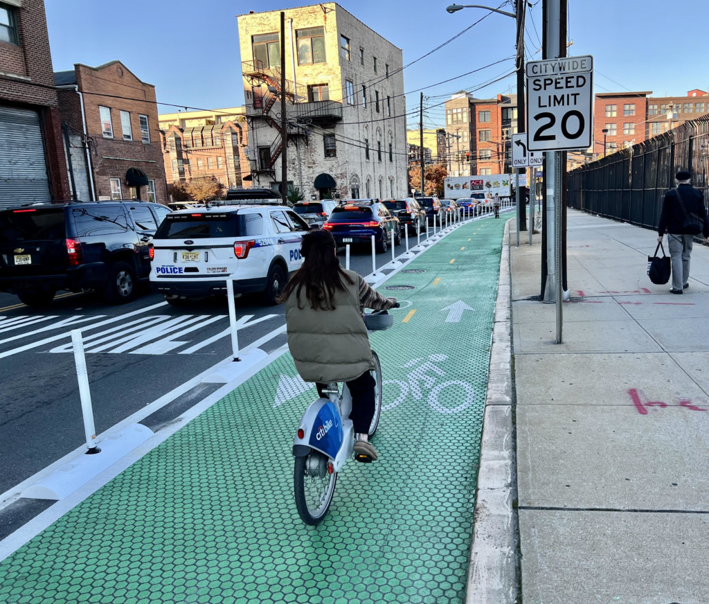 Person riding a bicycle on a green bike lane with cars on the street and buildings in the background.