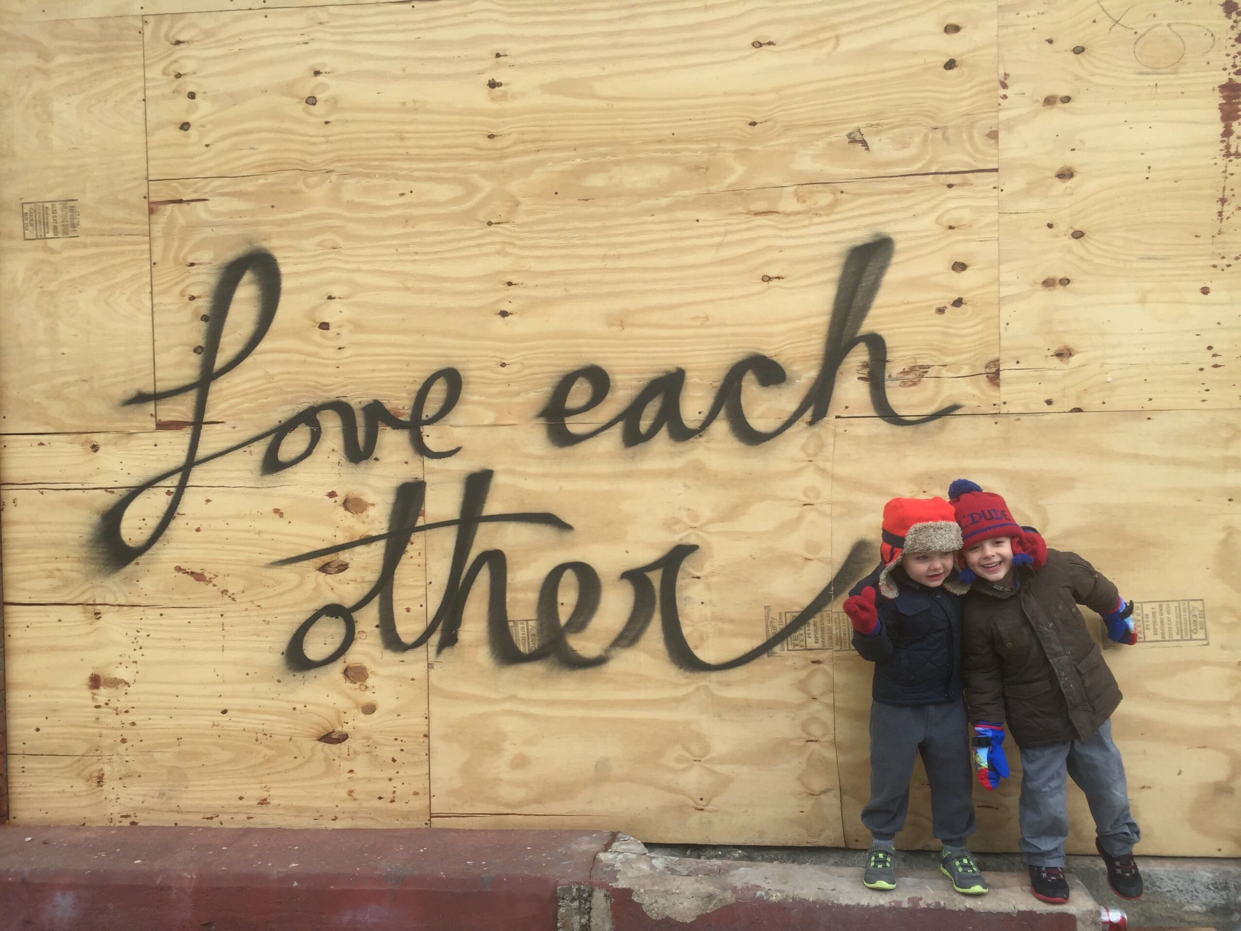 Two children stand in front of a wooden wall with the words 'Love each other' written on it, smiling and hugging.