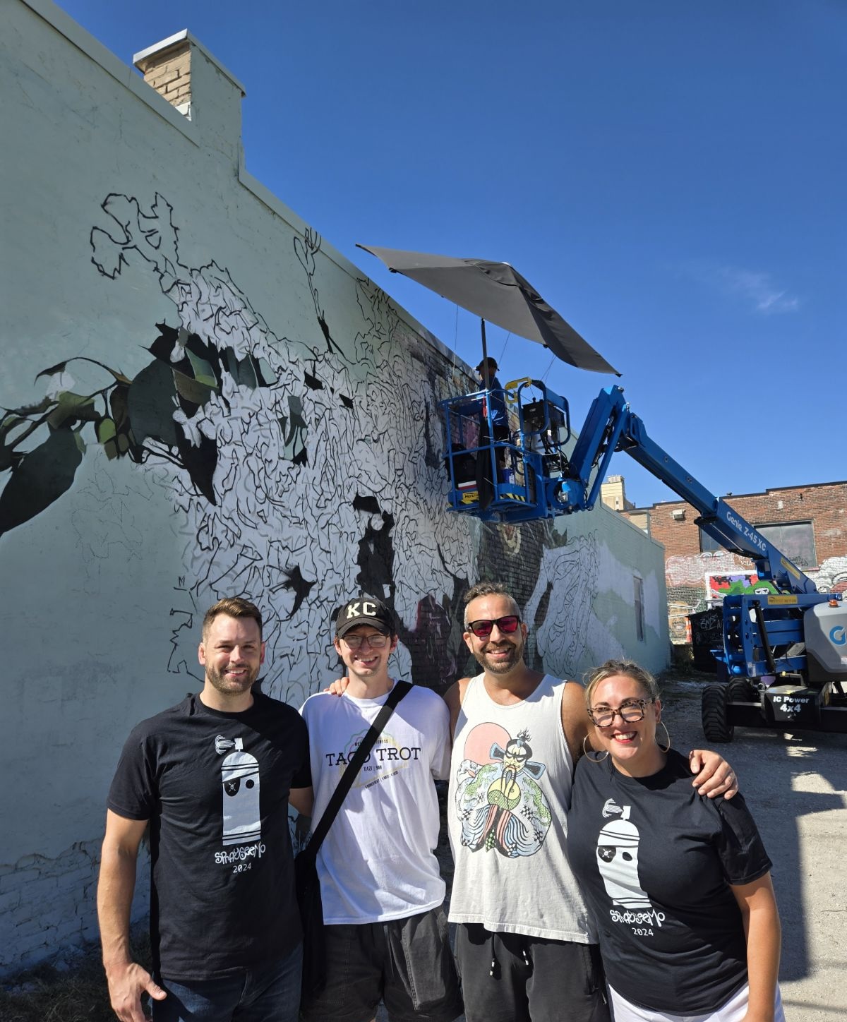 Four people stand in front of a mural being painted on a wall, with a lift and a worker painting above them.