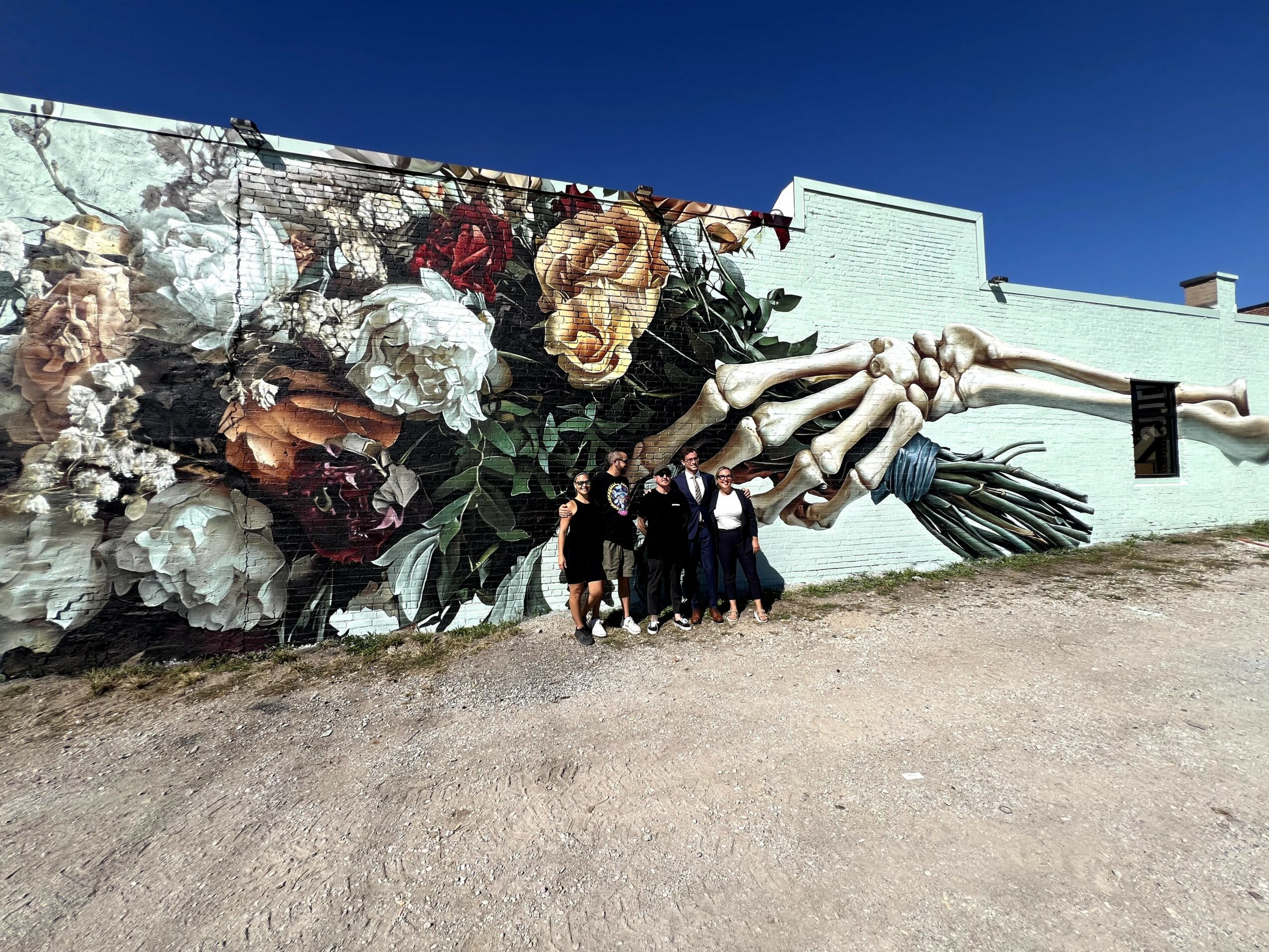 Group of five people standing in front of a large mural of flowers and a hand holding a broom, on a light blue wall.