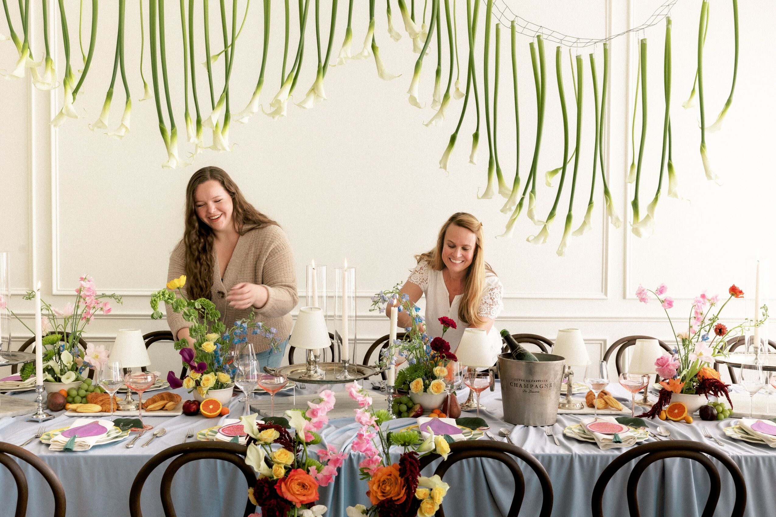 Two women standing behind a decorated table with flowers and food, smiling in a bright room with hanging green decorations.