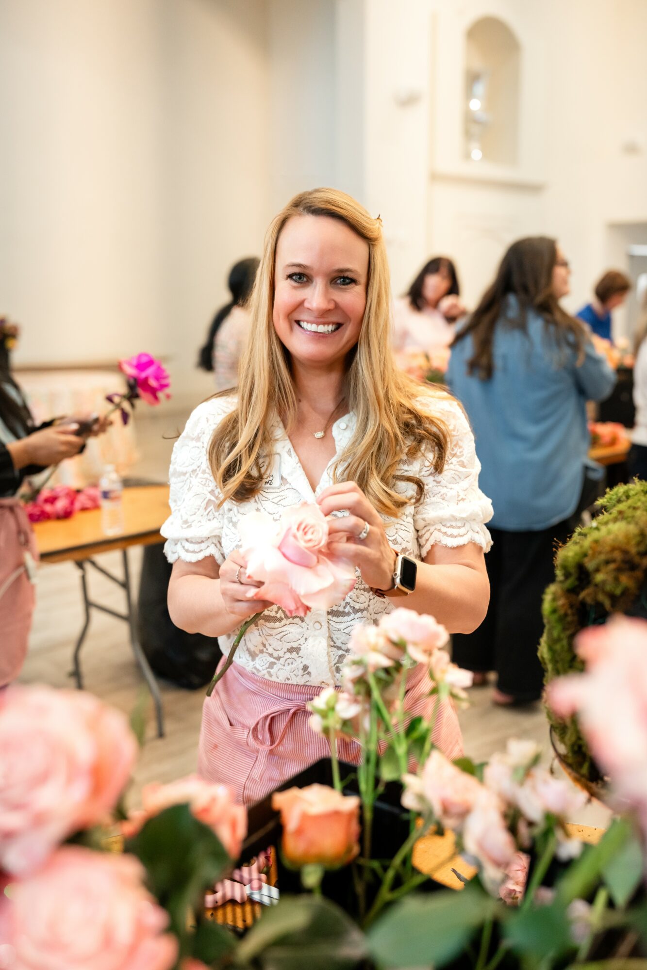 Smiling woman with long blonde hair in a white blouse and pink apron at a flower market, surrounded by flowers and people.
