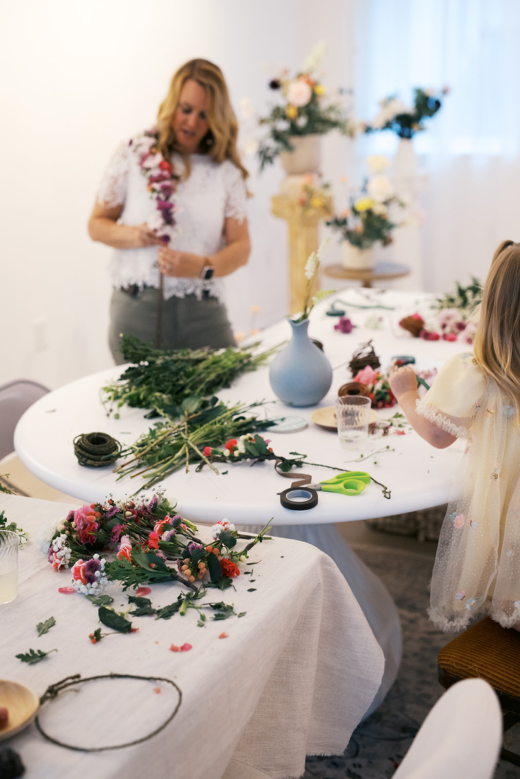 Woman and child making floral arrangements at a table with flowers, scissors, and greenery, in a bright room with vases and decorations.
