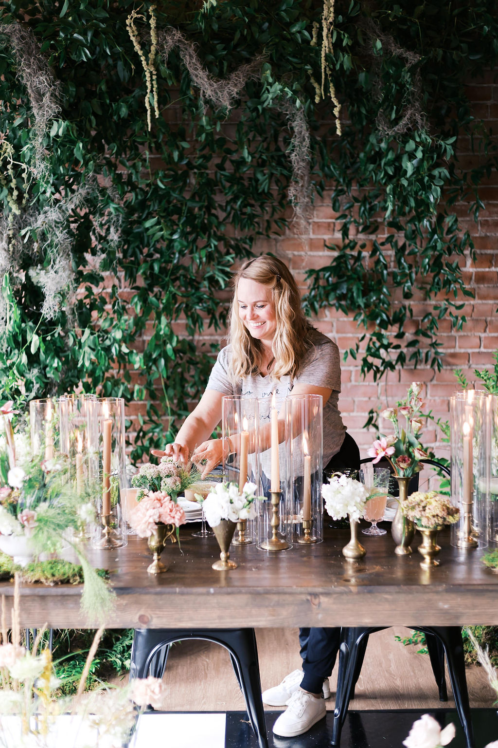 Woman arranging flowers at a decorated table outdoors with greenery and brick wall background.