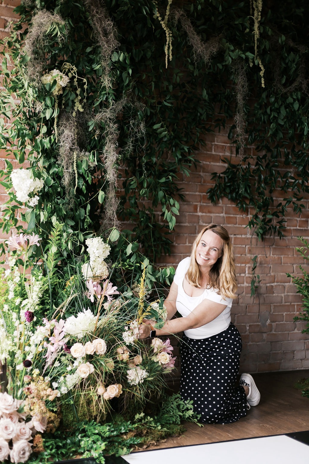 Young woman with long hair kneels beside a large floral arrangement against a brick wall, looking at the camera.