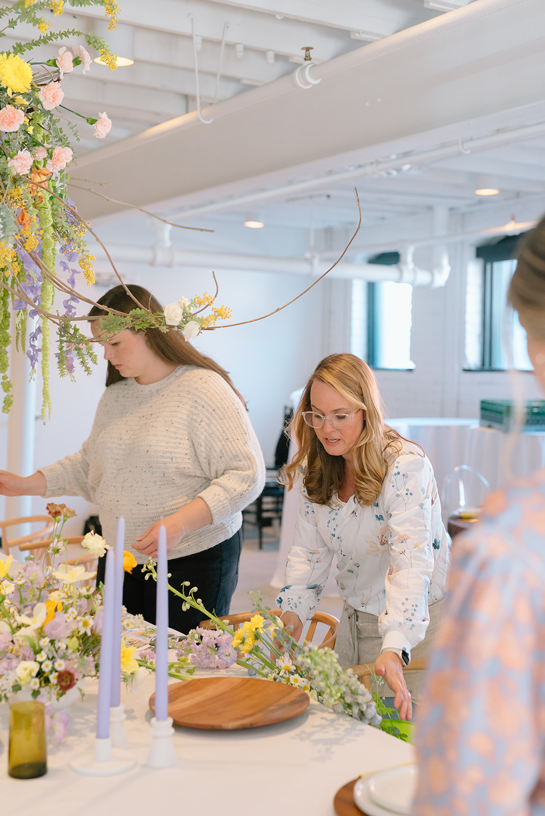 Two women arranging flowers at a table with candles and floral decorations in a bright, modern room.