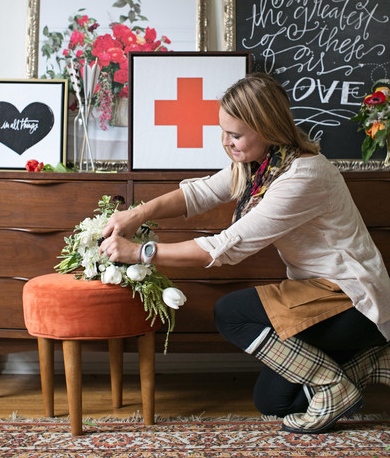 Woman arranging flowers on a small orange stool in a room with framed artwork and flowers on a wooden surface.