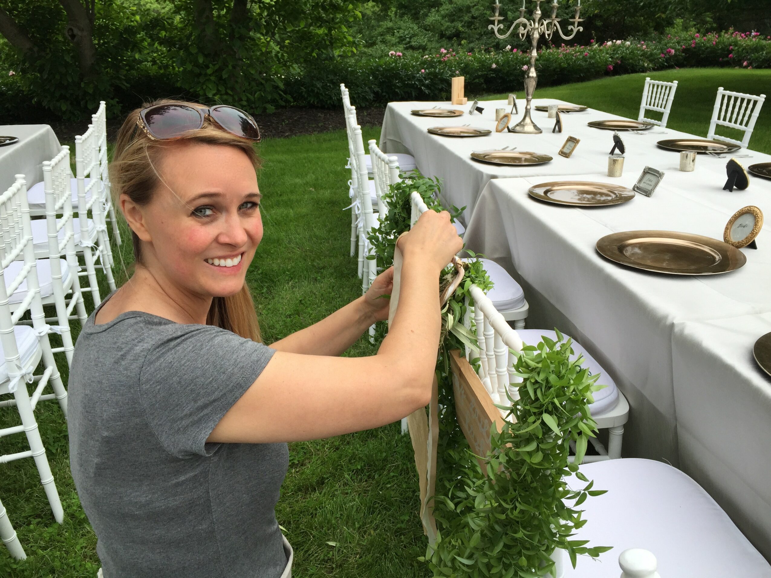Woman smiling and decorating outdoor table with greenery, set for a formal event, in a garden setting.