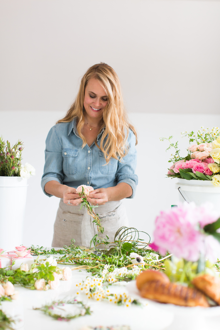 Woman arranging flowers at a table with various flowers and greenery, smiling, in a bright room.