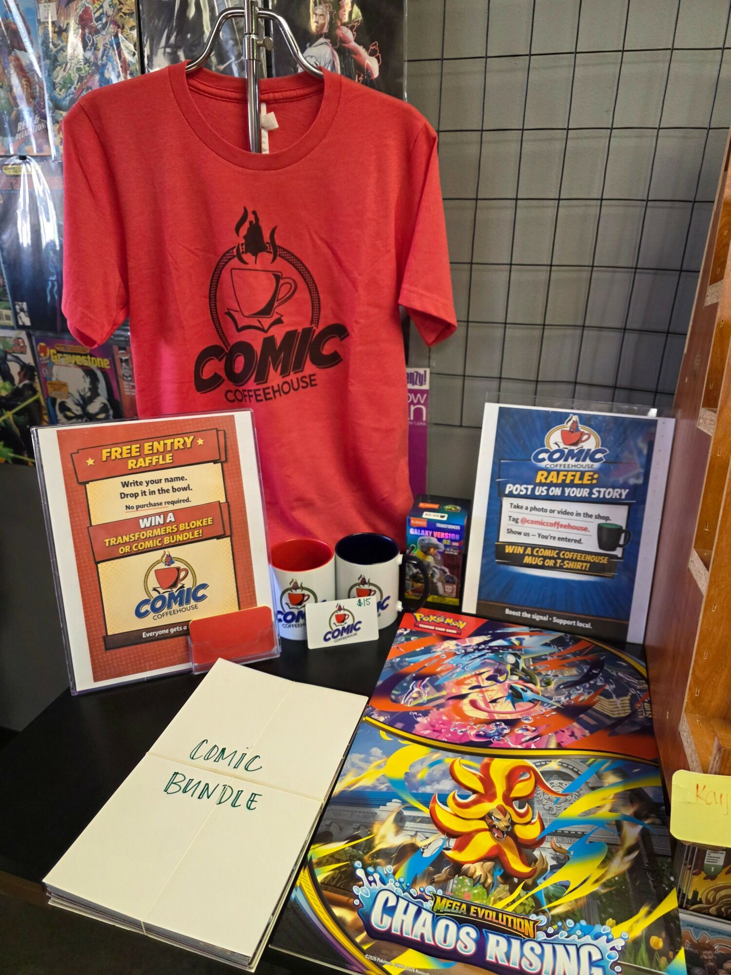 Red T-shirt with a comic book logo displayed on a table with comic books and promotional materials.