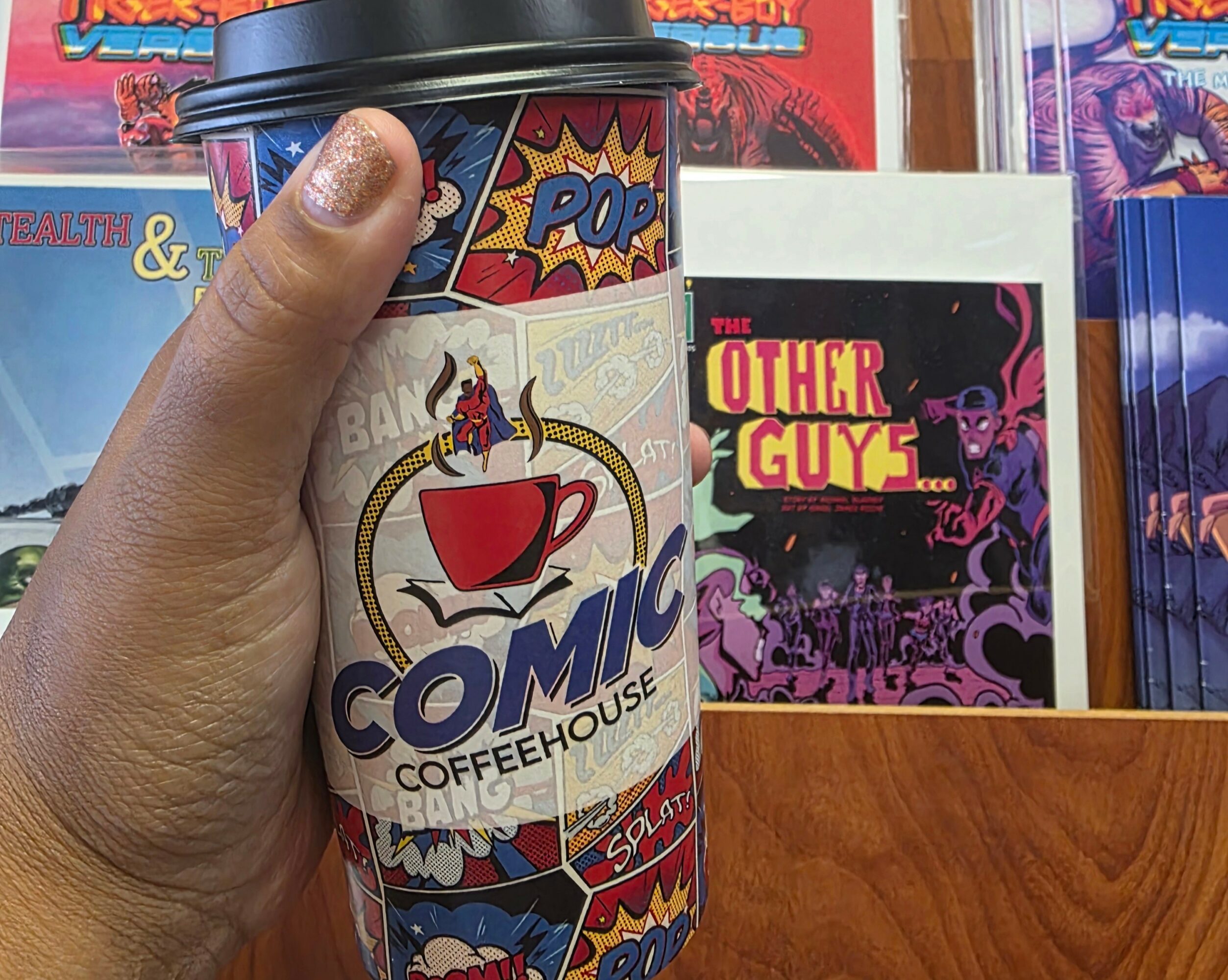 Hand holding a comic-themed coffee cup with colorful comic book graphics, in front of comic books on a wooden surface.