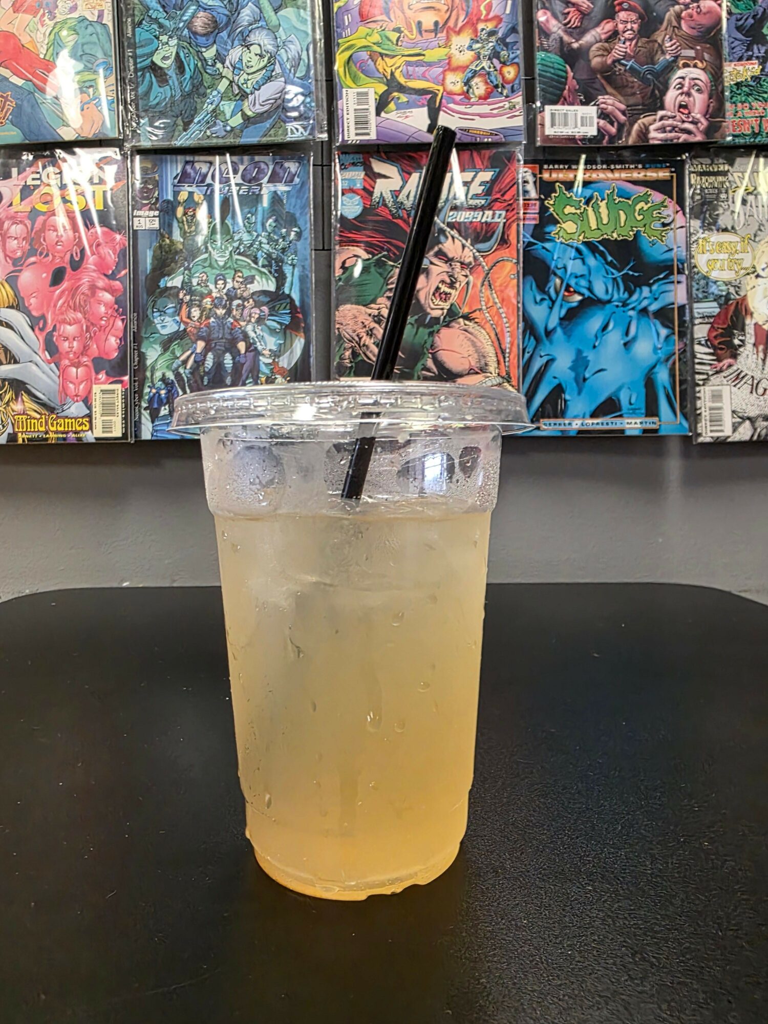 A large iced beverage in a clear plastic cup with a black straw, placed on a black surface, with comic books in the background.