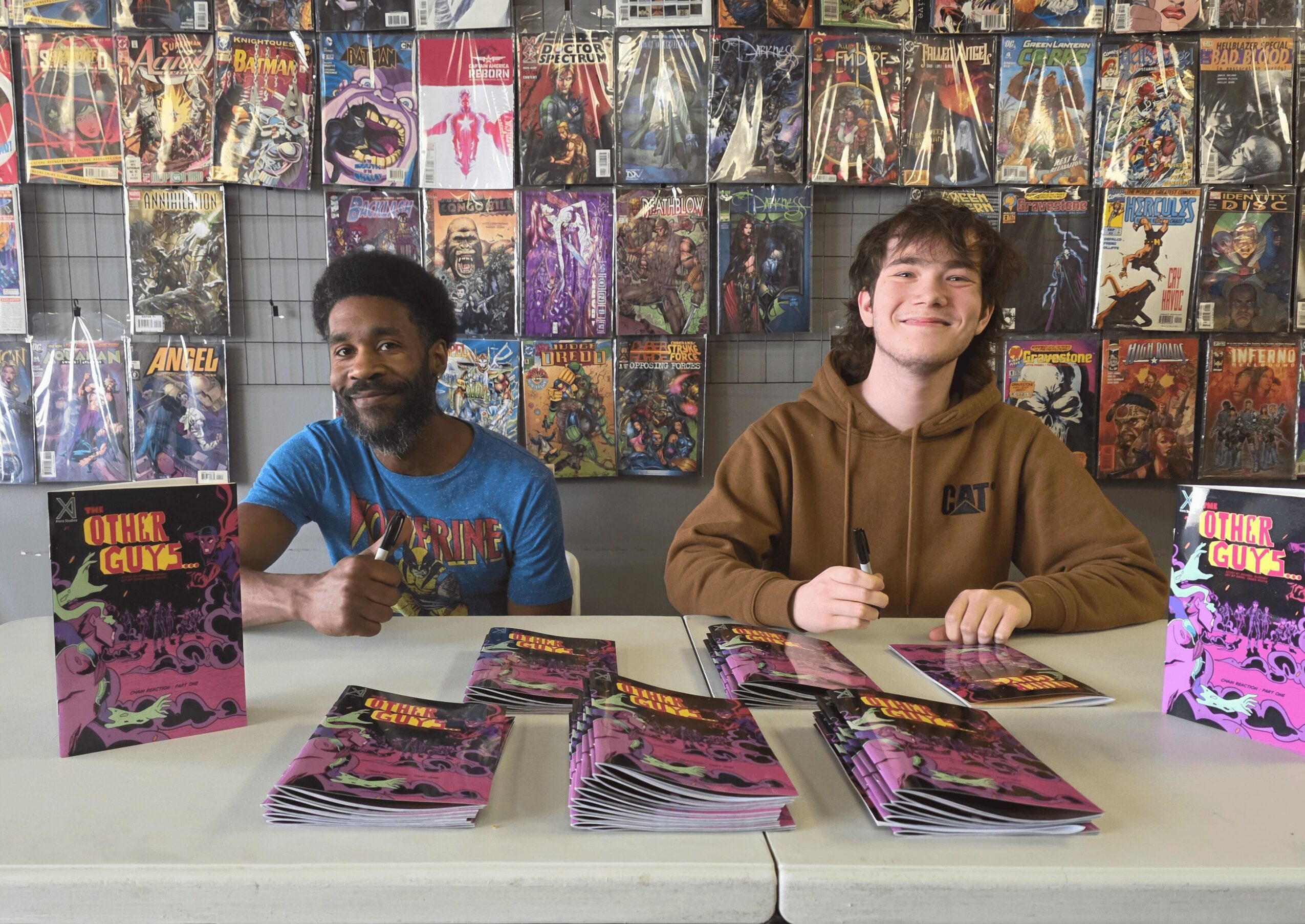Two people sit at a table with comic books, smiling, in front of a wall of comic book covers, one with a brown hoodie, the other with a blue shirt.