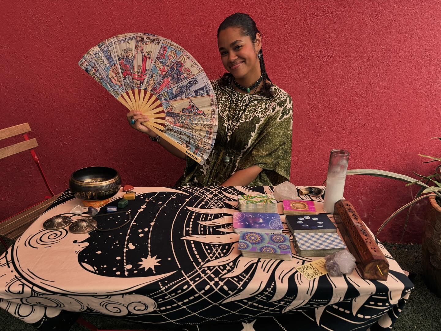 Woman holding a decorative fan, sitting at a table with a patterned cloth, against a pink wall.