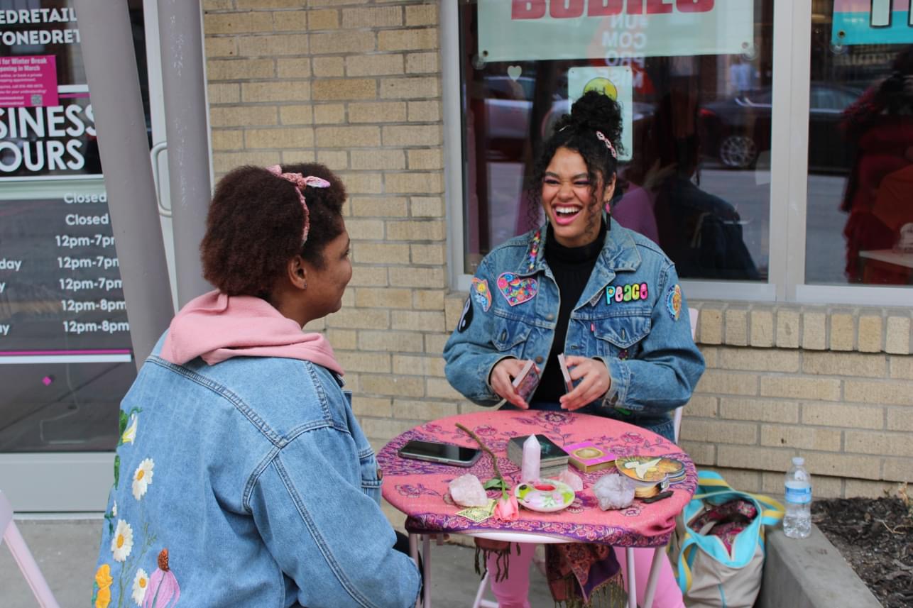 Two women sit at a pink table outside a store, smiling and talking, with food and drinks on the table.