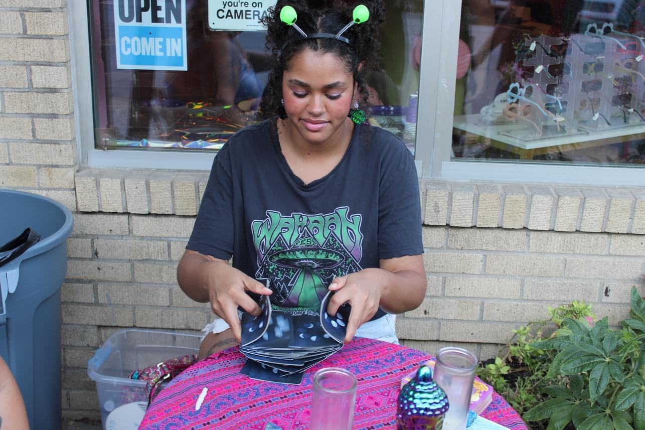 Girl wearing headband with green antennae, sitting outside with pink blanket, holding a small bag, in front of a brick wall and window.