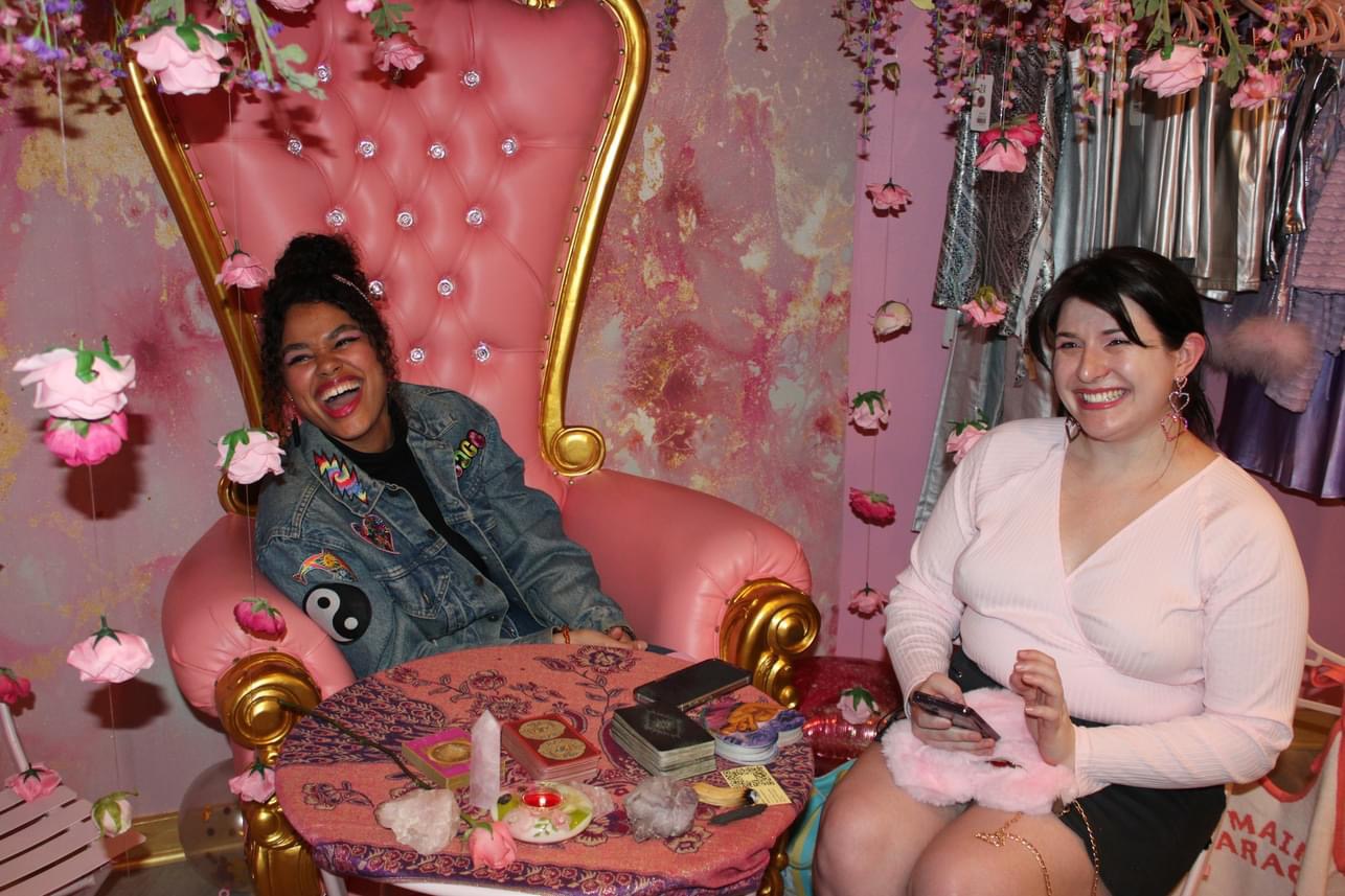 Two women smiling, one sitting on a pink ornate chair, the other standing nearby, in a decorated room with pink flowers and curtains.