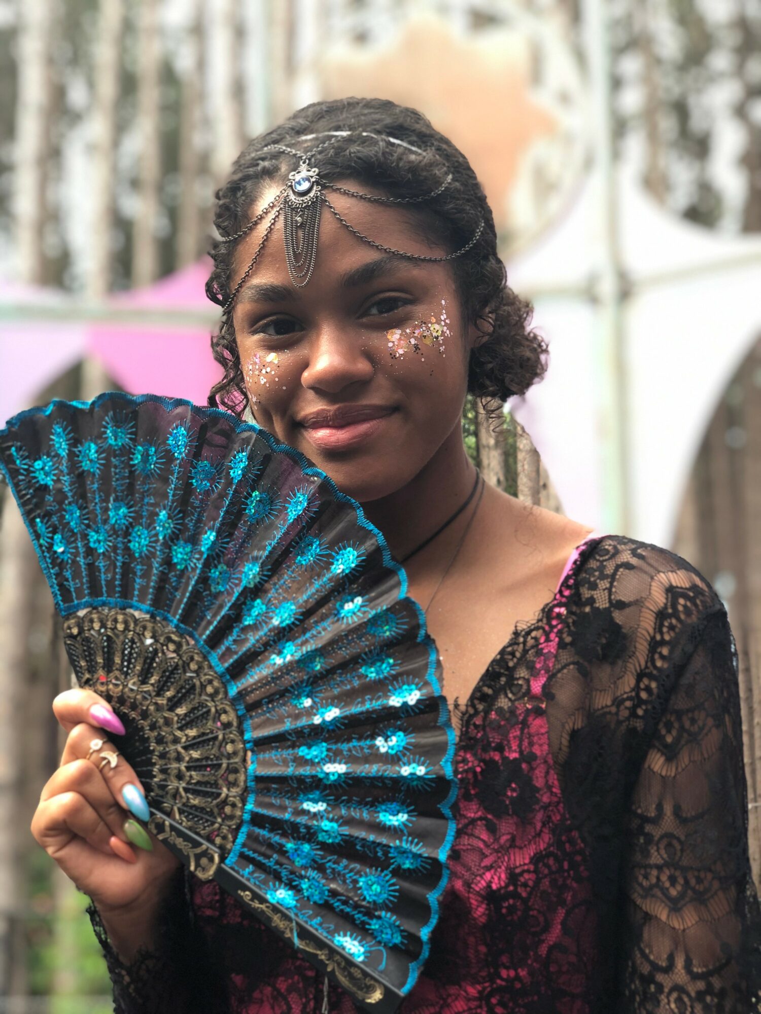 Young woman with dark hair and jewelry holding a blue and black fan, wearing a lace dress, outdoors.