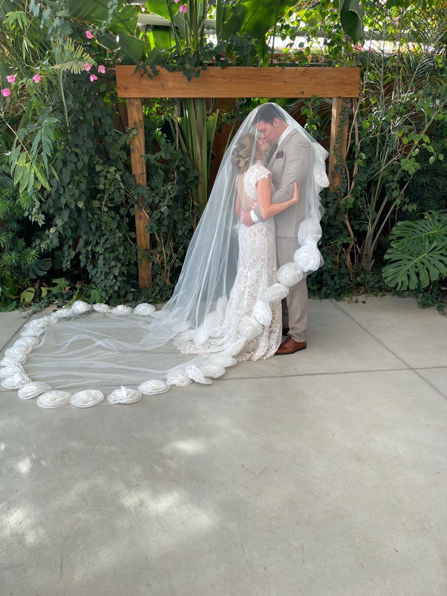 Person in a wedding dress with a long veil, standing outdoors on a concrete surface, surrounded by greenery and flowers.