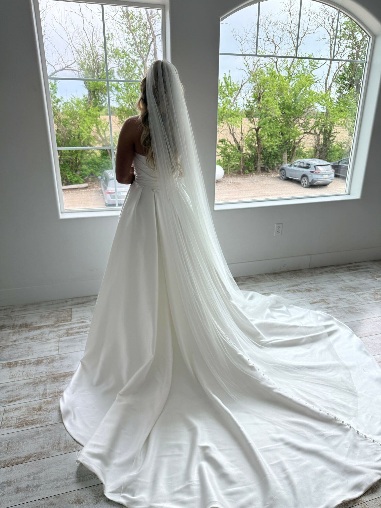 Person in a white dress standing near a window with trees outside, sunlight streaming in, and a bed in the room.