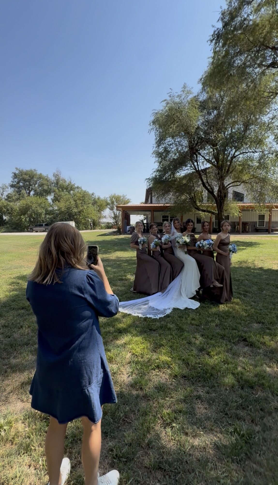 Person in blue dress taking photo of a wedding party outdoors under a tree on a sunny day.
