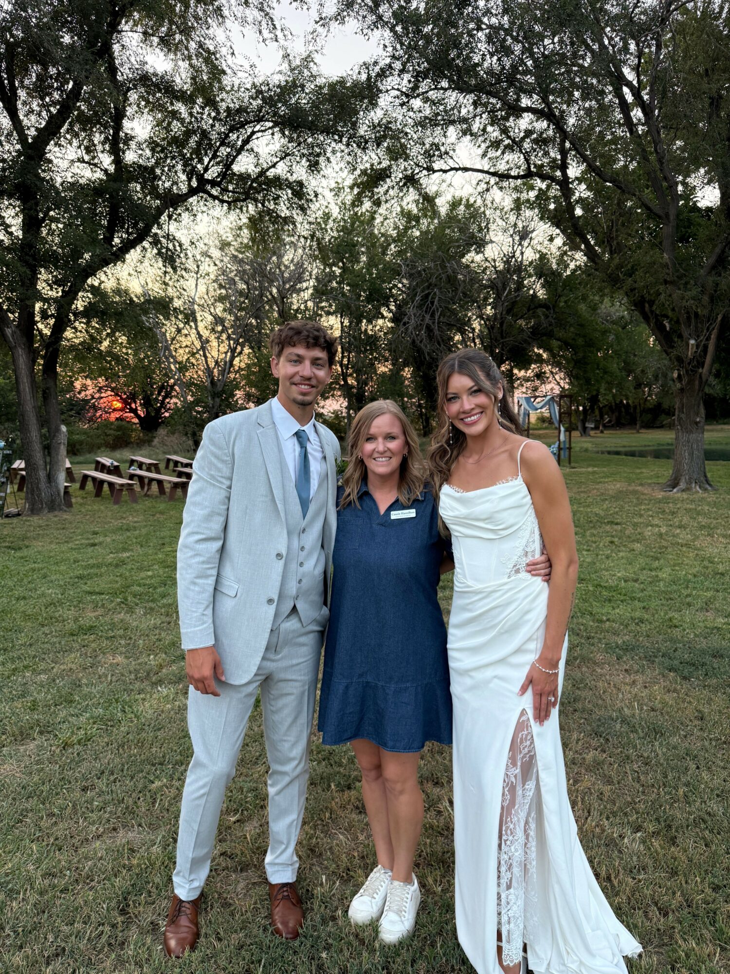 Three people standing outdoors on grass, with trees and a sunset in the background.