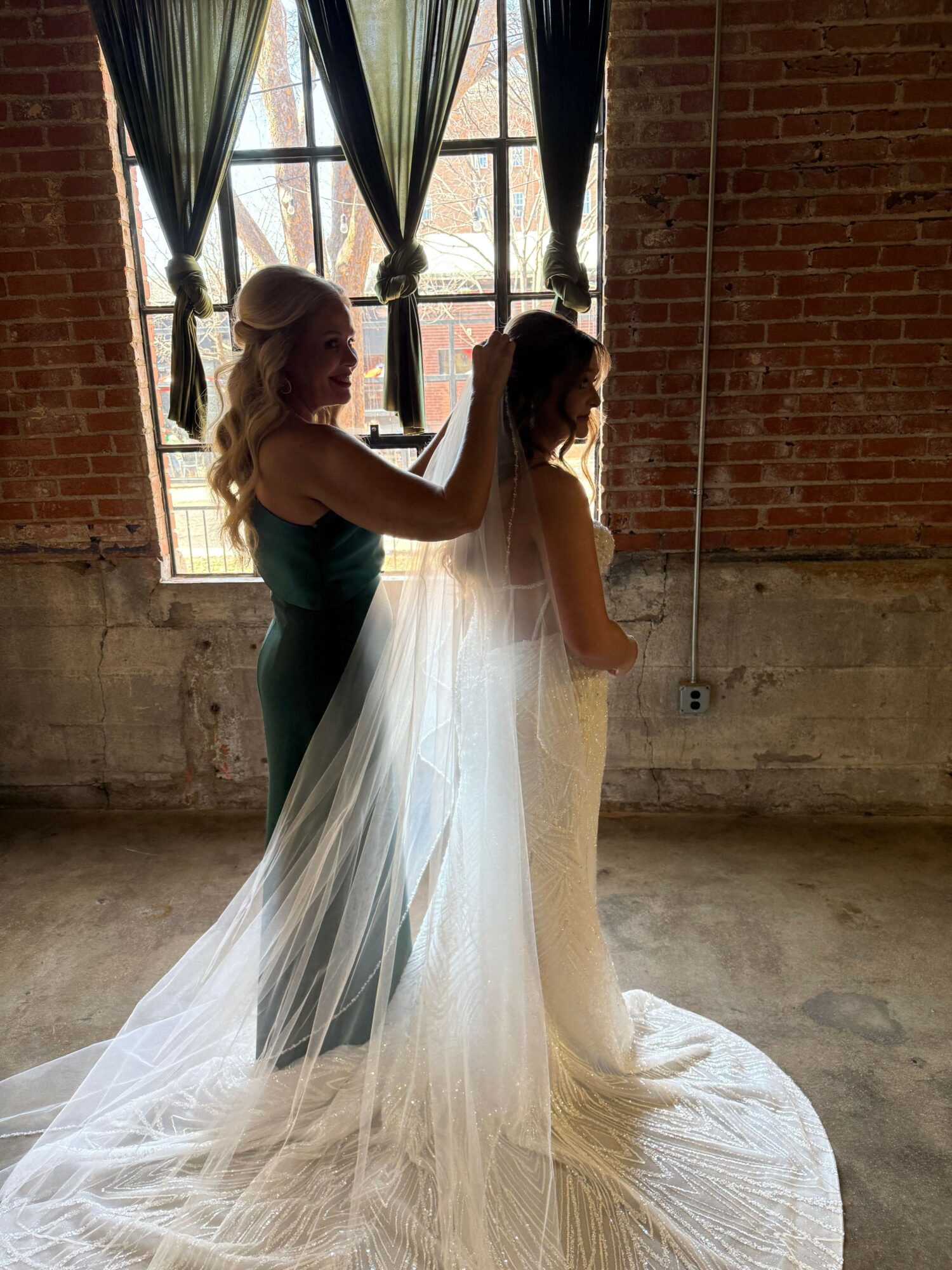 A bride in a wedding dress stands indoors near a window with brick walls, holding a bouquet, facing sideways.