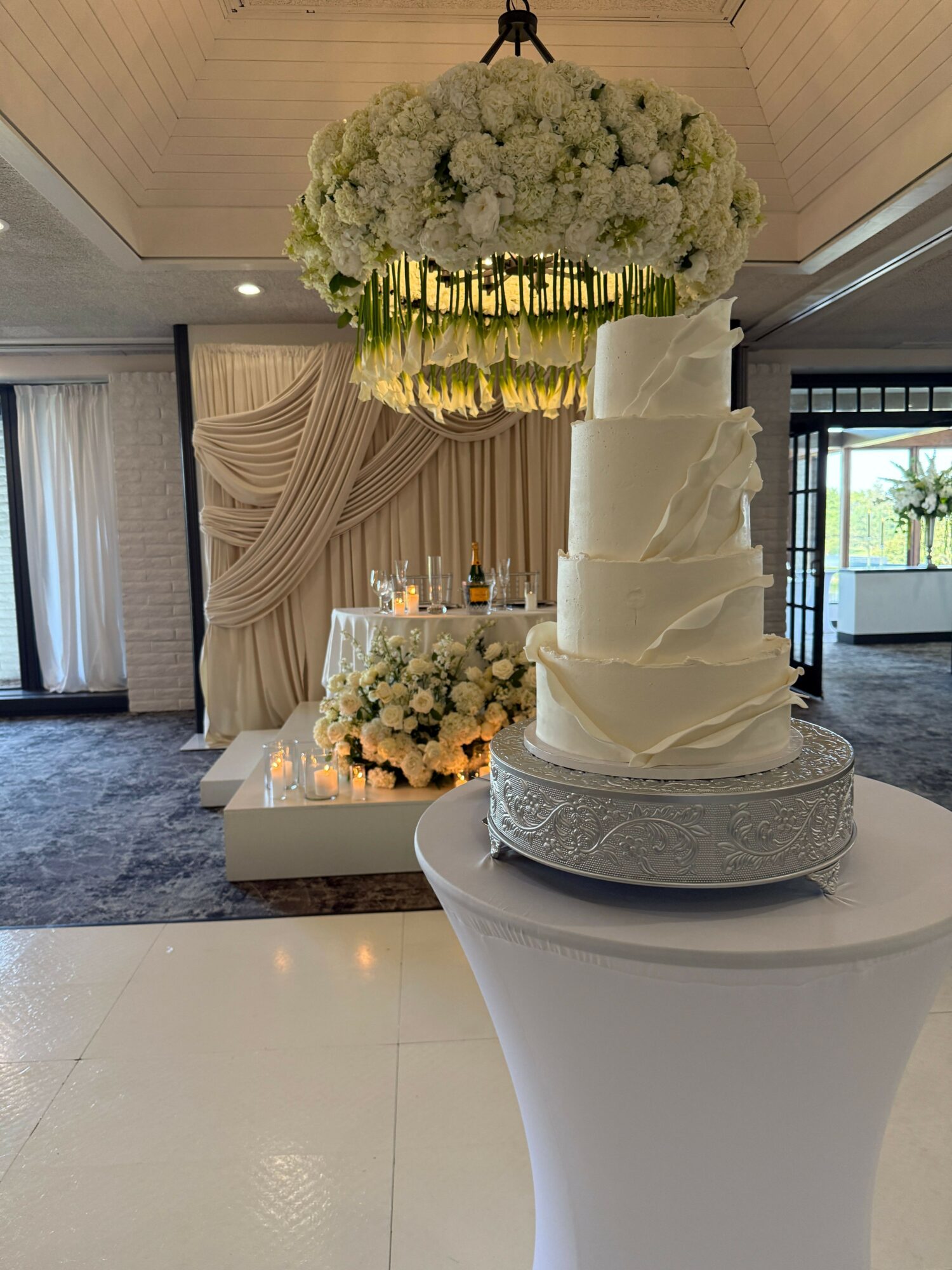Wedding cake with white flowers and candles on a table, with a bouquet of white flowers nearby, in a bright room.
